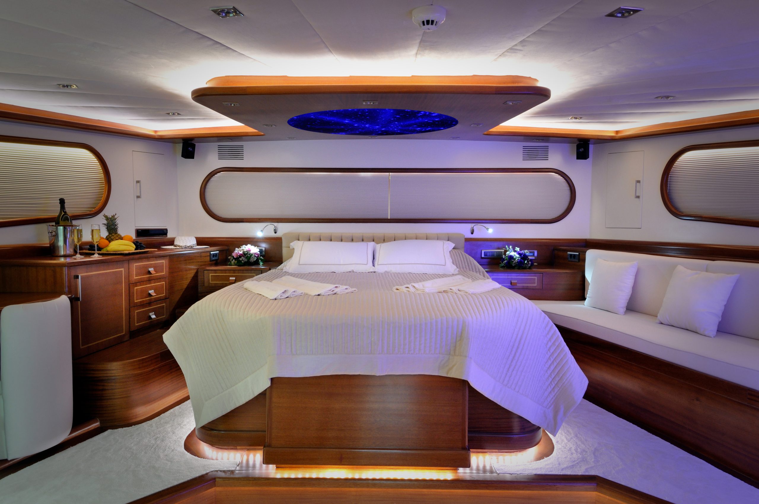 DOLCE MARE A luxurious yacht bedroom with a large bed, wooden furniture, soft lighting, a curved white sofa, decorative pillows, and a ceiling panel with a starry sky design. Wine and fruit sit on a side cabinet.