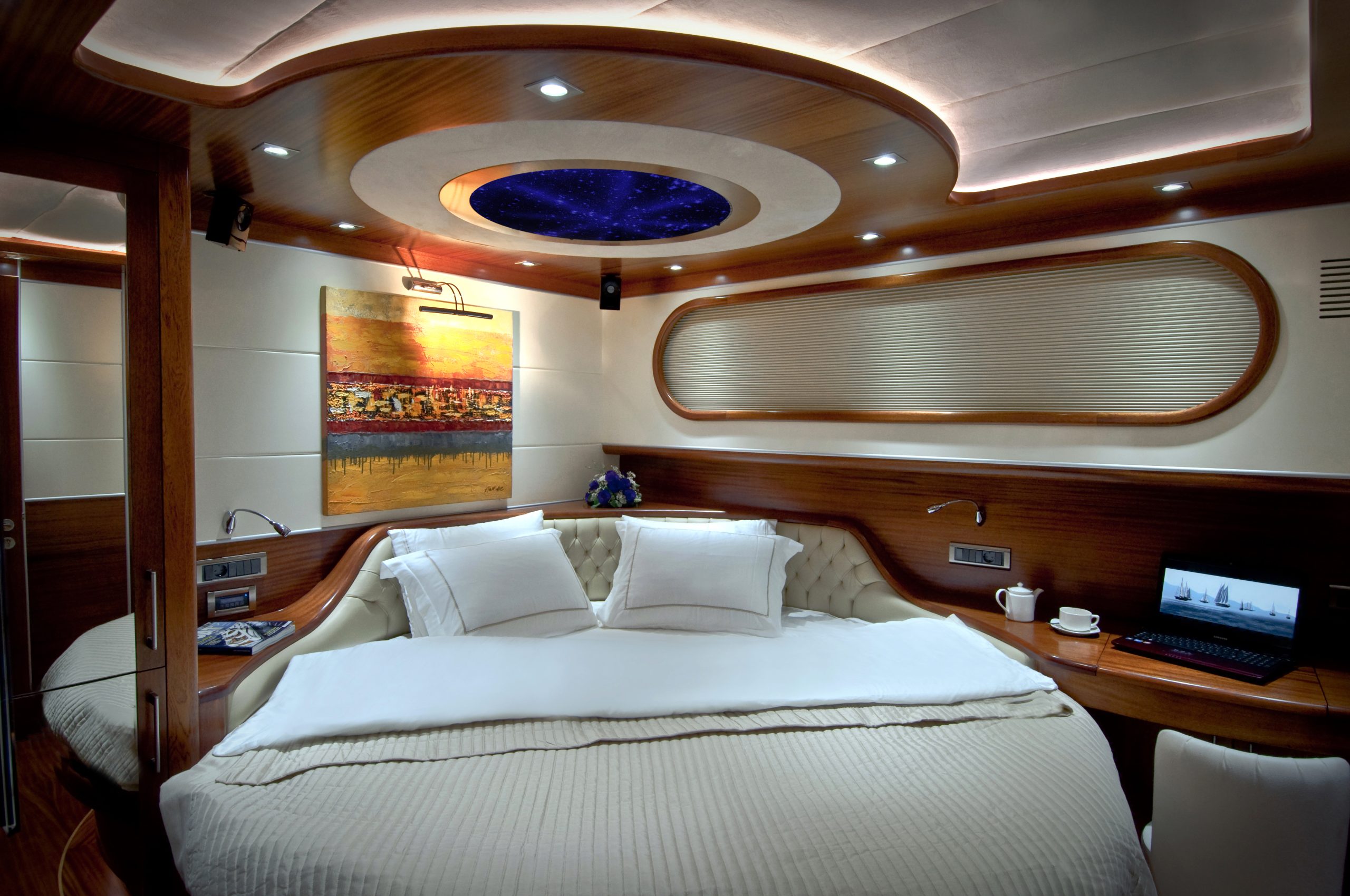 DOLCE MARE A modern yacht cabin with a large bed, white bedding, wood accents, abstract wall art, a built-in desk with a laptop, and soft ambient lighting including a ceiling feature resembling a starry sky.