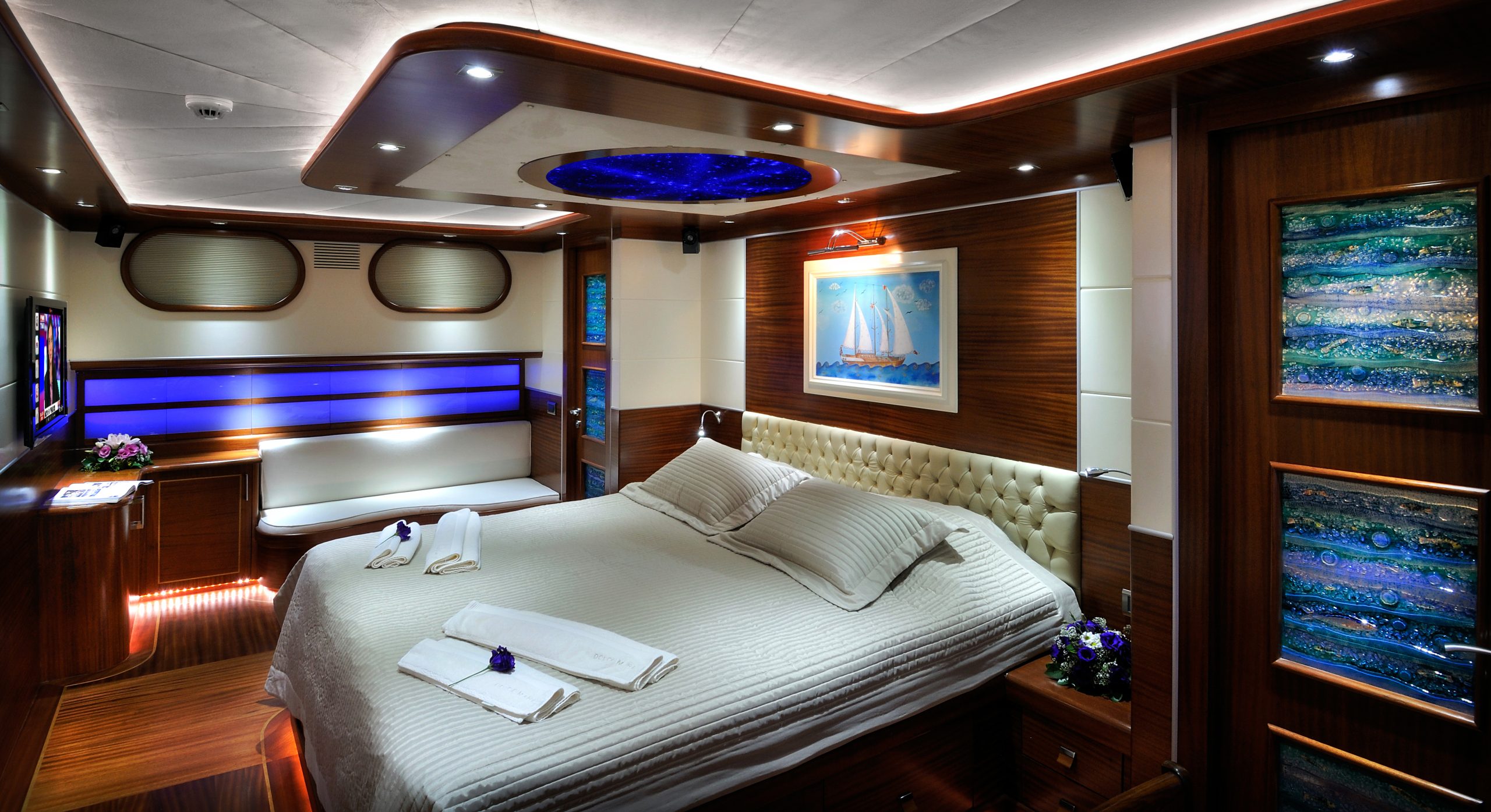 DOLCE MARE A luxurious yacht bedroom with a large bed, elegant wood paneling, blue accent lighting, modern art, and decorative details, including folded towels and flowers on the bed and side surfaces.
