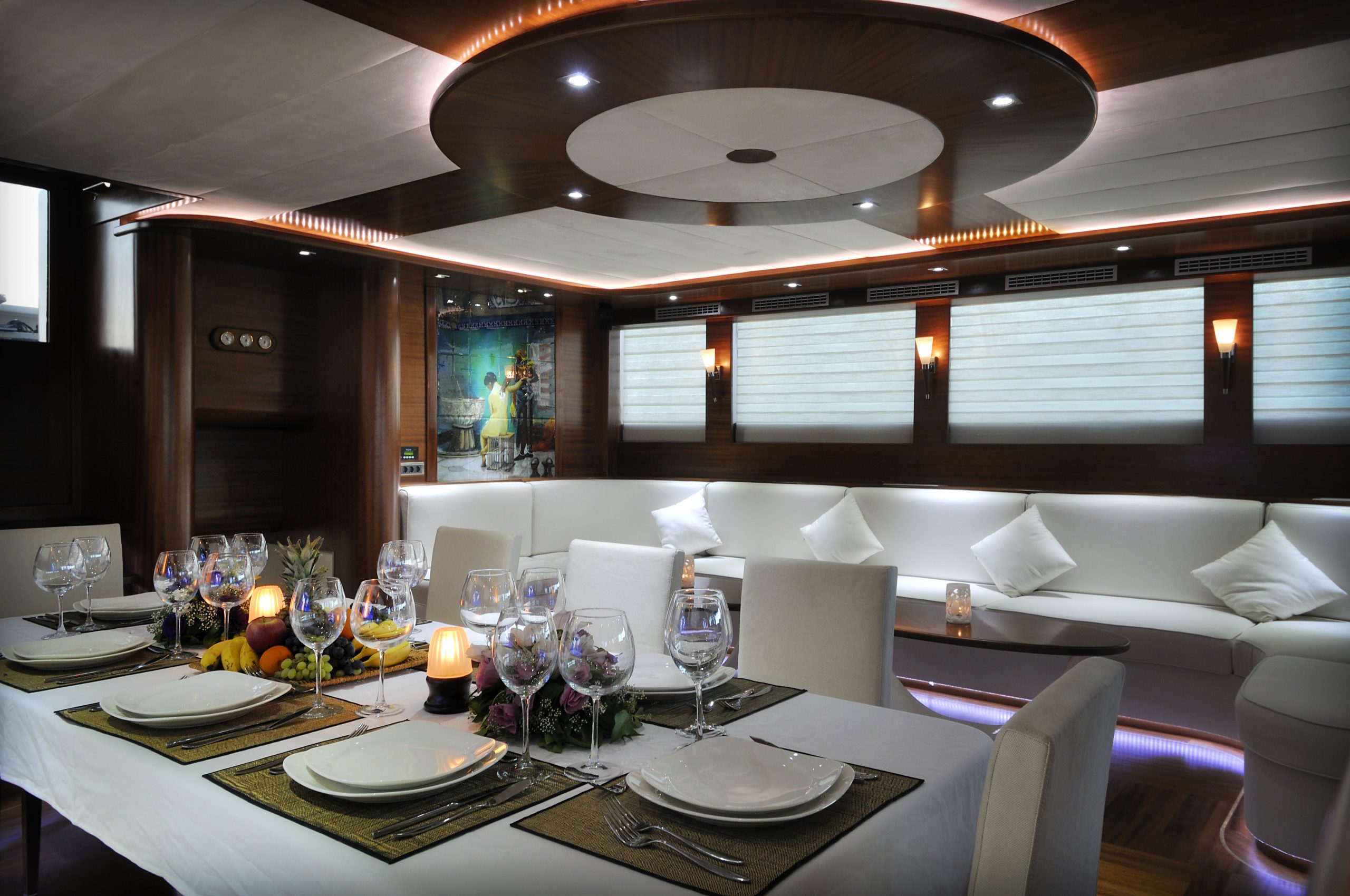 DOLCE MARE A luxurious yacht interior with a modern design, featuring a dining table set for four with elegant place settings, wine glasses, and flowers. White sofas line the wall, and soft lighting creates a warm ambiance.