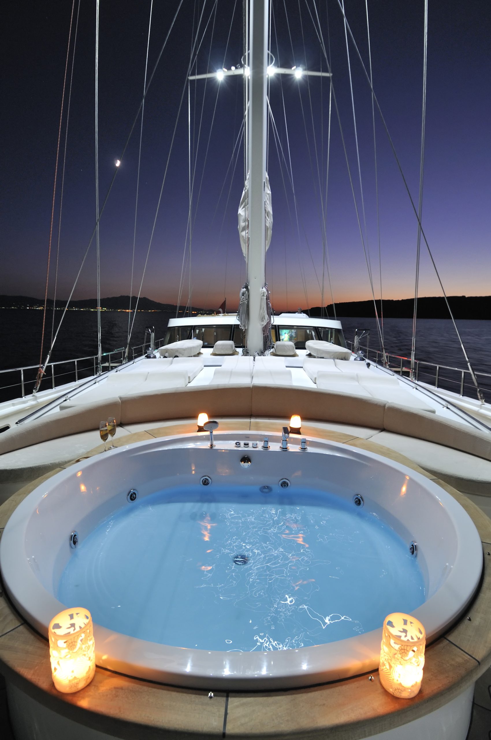 DOLCE MARE A luxury yacht deck at sunset features a round jacuzzi with glowing candles on the edge, white lounge seating in the background, and calm waters surrounding the vessel.