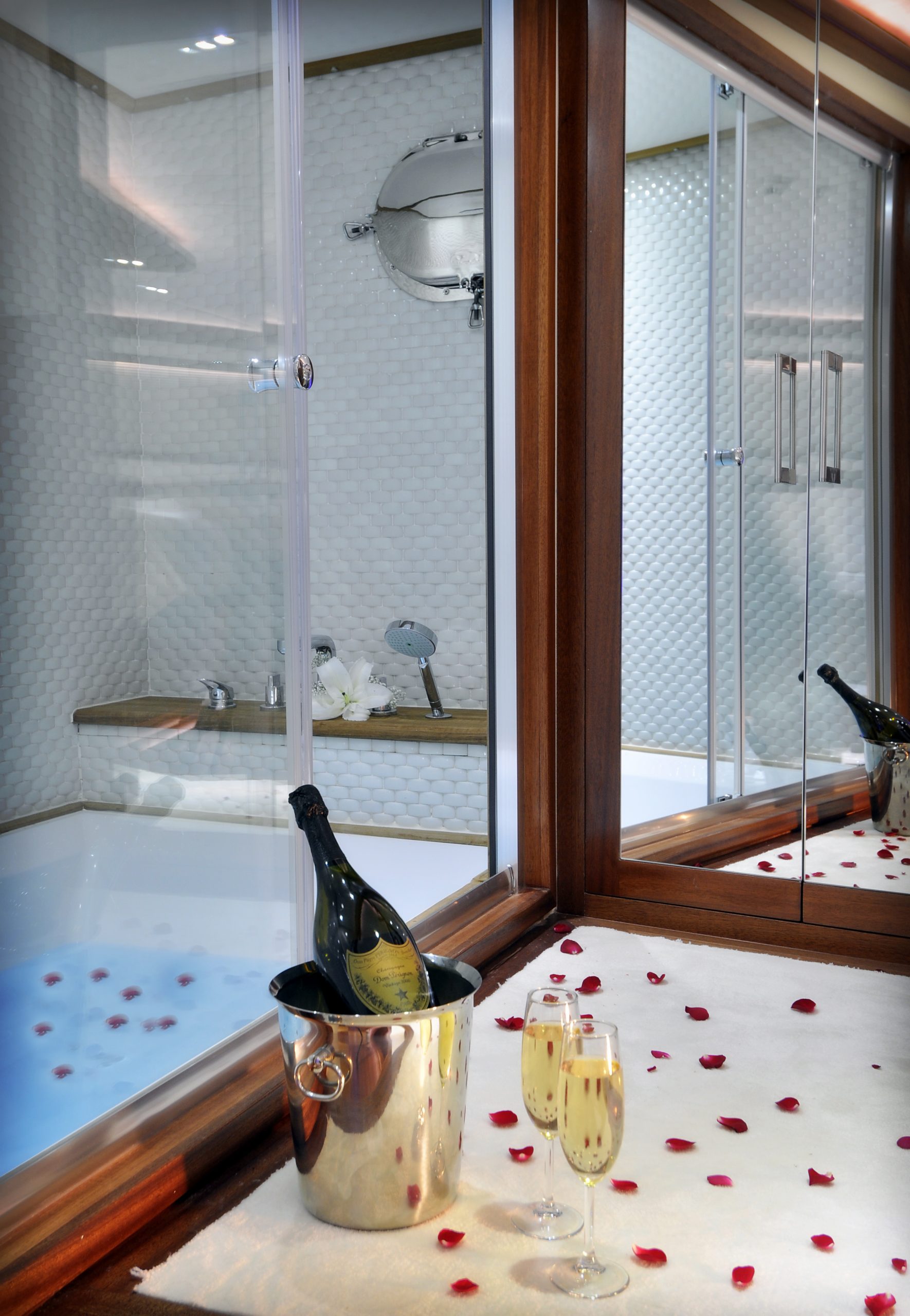 DOLCE MARE A luxurious bathroom with a bathtub, rose petals scattered on a white towel by the tub, a bottle of champagne in an ice bucket, and two filled champagne glasses, with a large mirror reflecting the scene.