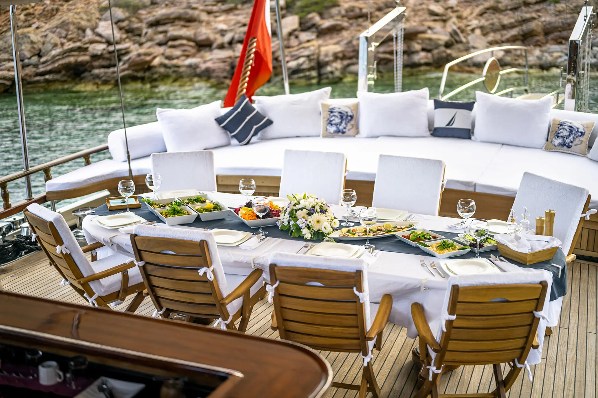 An elegant outdoor dining table set with plates, glasses, flowers, and food on a yacht deck, with cushioned seating in the background overlooking water and rocky shoreline.