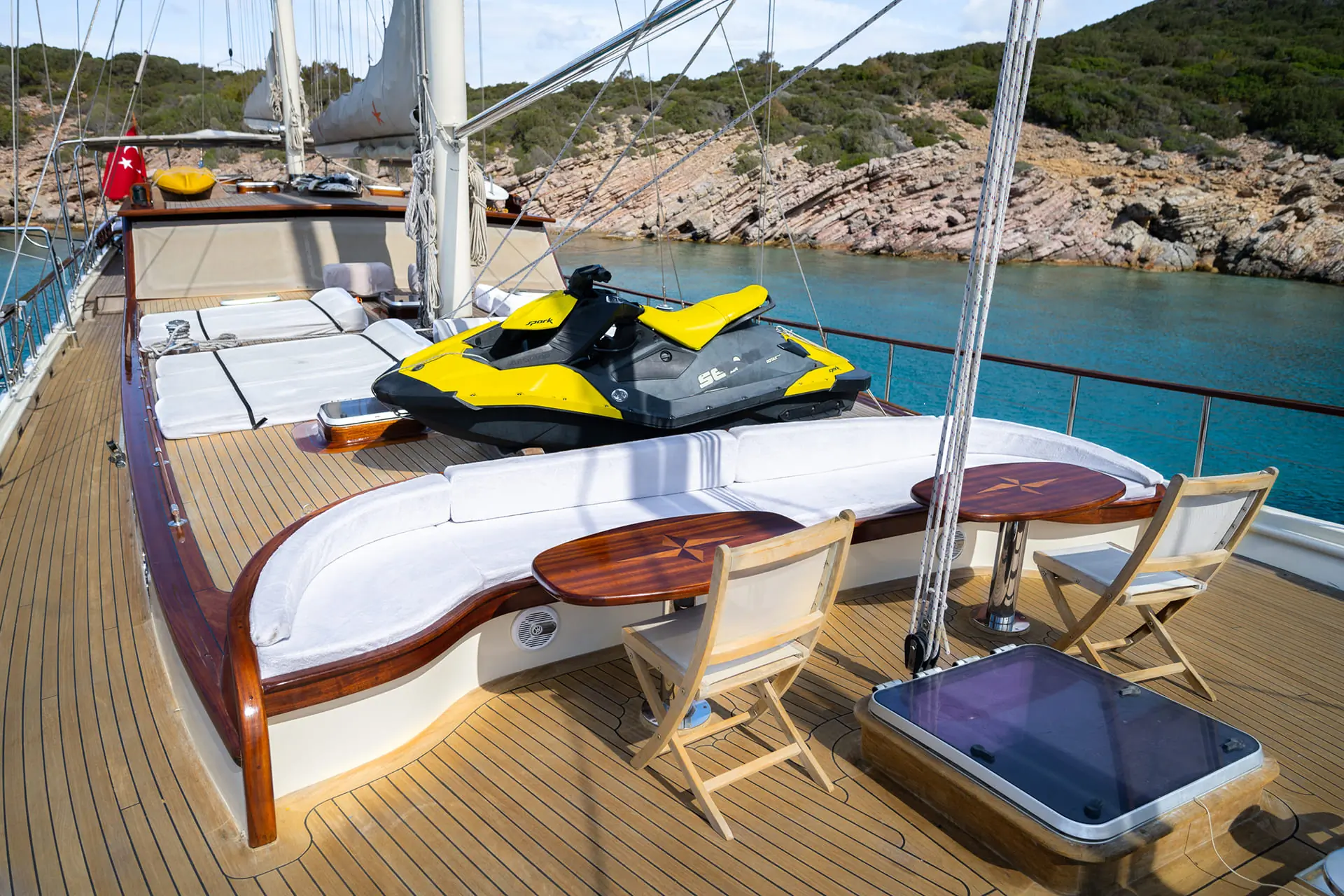 A yacht deck with cushioned seating, wooden tables, chairs, and a yellow jet ski. Turquoise water and rocky hills are visible in the background.