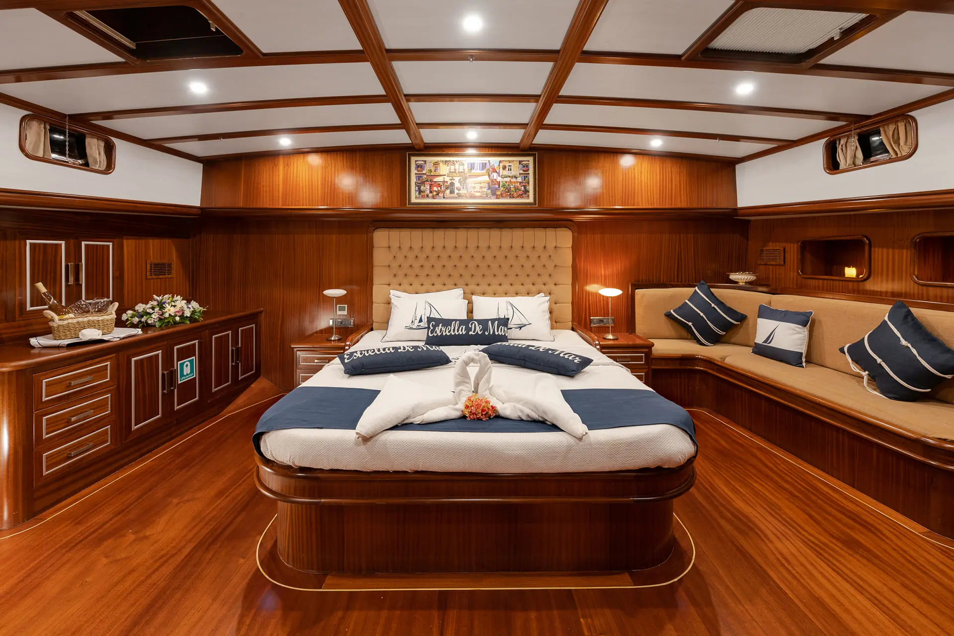 Luxurious yacht cabin with polished wood, a large bed with white and navy pillows, a cushioned bench, flowers, and warm lighting, creating an elegant and inviting atmosphere.