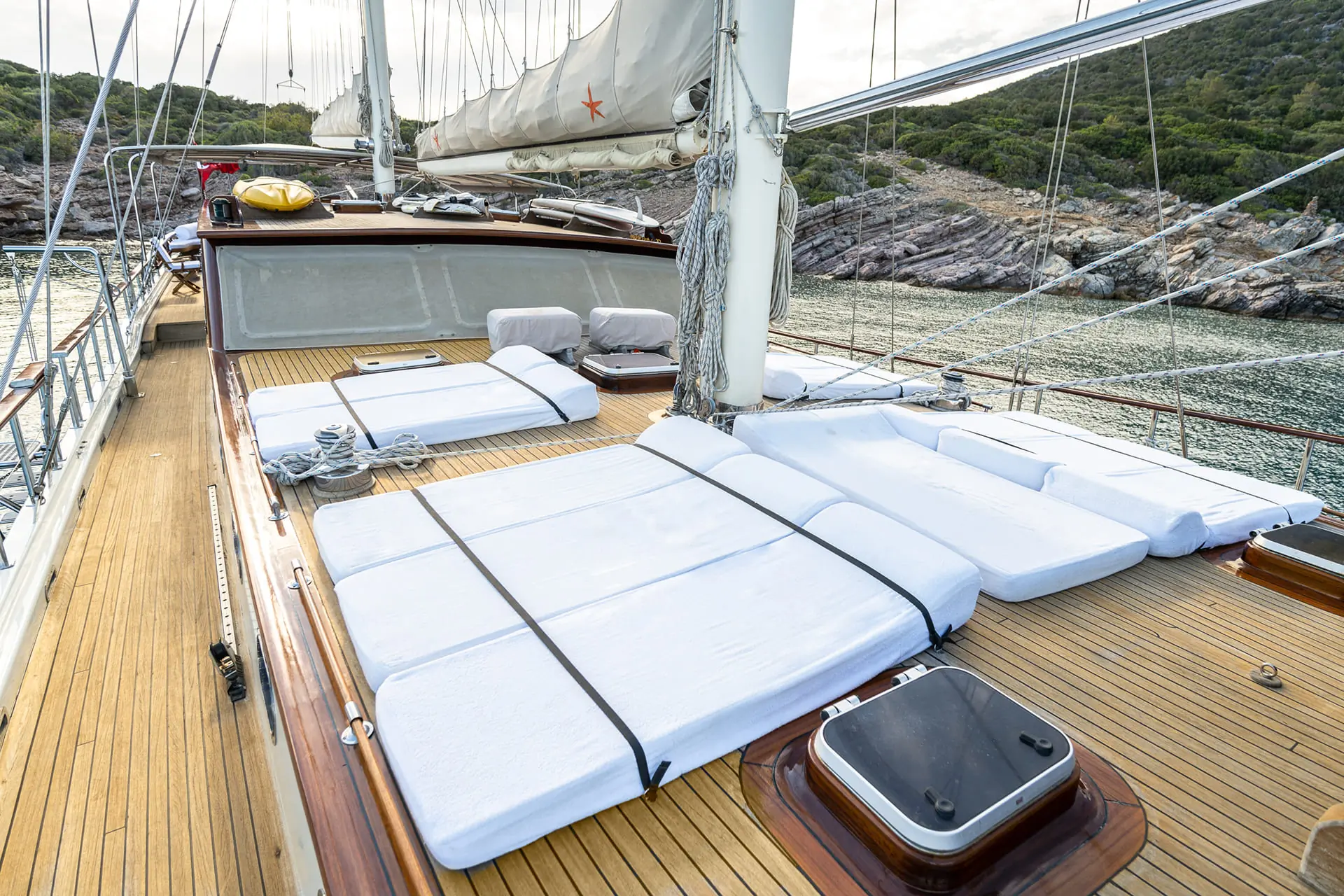The deck of a sailboat with white cushioned sunbeds, ropes, and sails, surrounded by rocky shoreline and green hills in the background.