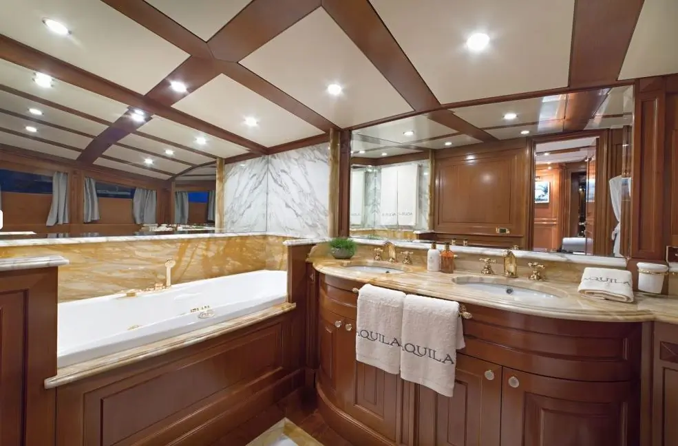 Luxurious bathroom with wooden cabinets, marble countertops, a large whirlpool tub, double sink, and soft lighting. Towels labeled AQUILA are neatly placed beside the sinks.