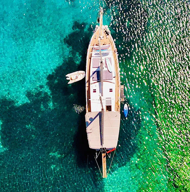 Aerial view of a sailboat anchored in clear, turquoise water with a small dinghy nearby. Sunlight sparkles on the water, revealing varying shades of blue and green beneath the boat.
