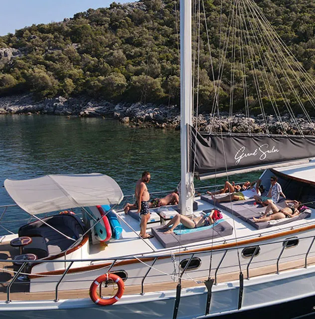 People relax and sunbathe on the deck of a sailboat anchored near a lush, green shoreline with calm water and trees in the background.