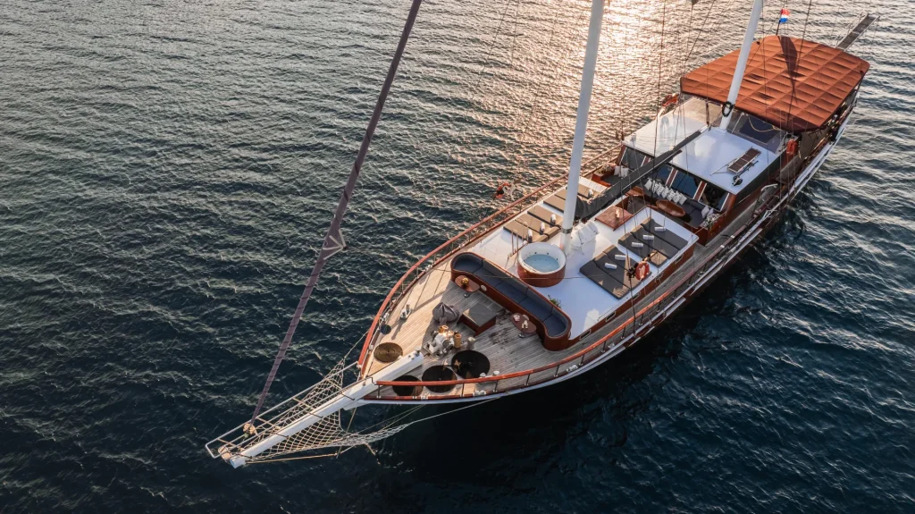 Aerial view of a luxurious yacht with a hot tub on deck, anchored in calm, blue water under the sunlight. The yacht features wooden accents and shaded seating areas.