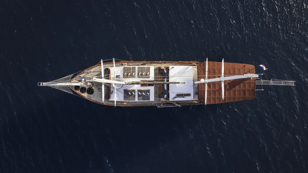 Aerial view of a sailboat with white deck and brown seating areas, floating on dark blue water. The boat’s masts and rigging are visible, along with a small flag at the stern.