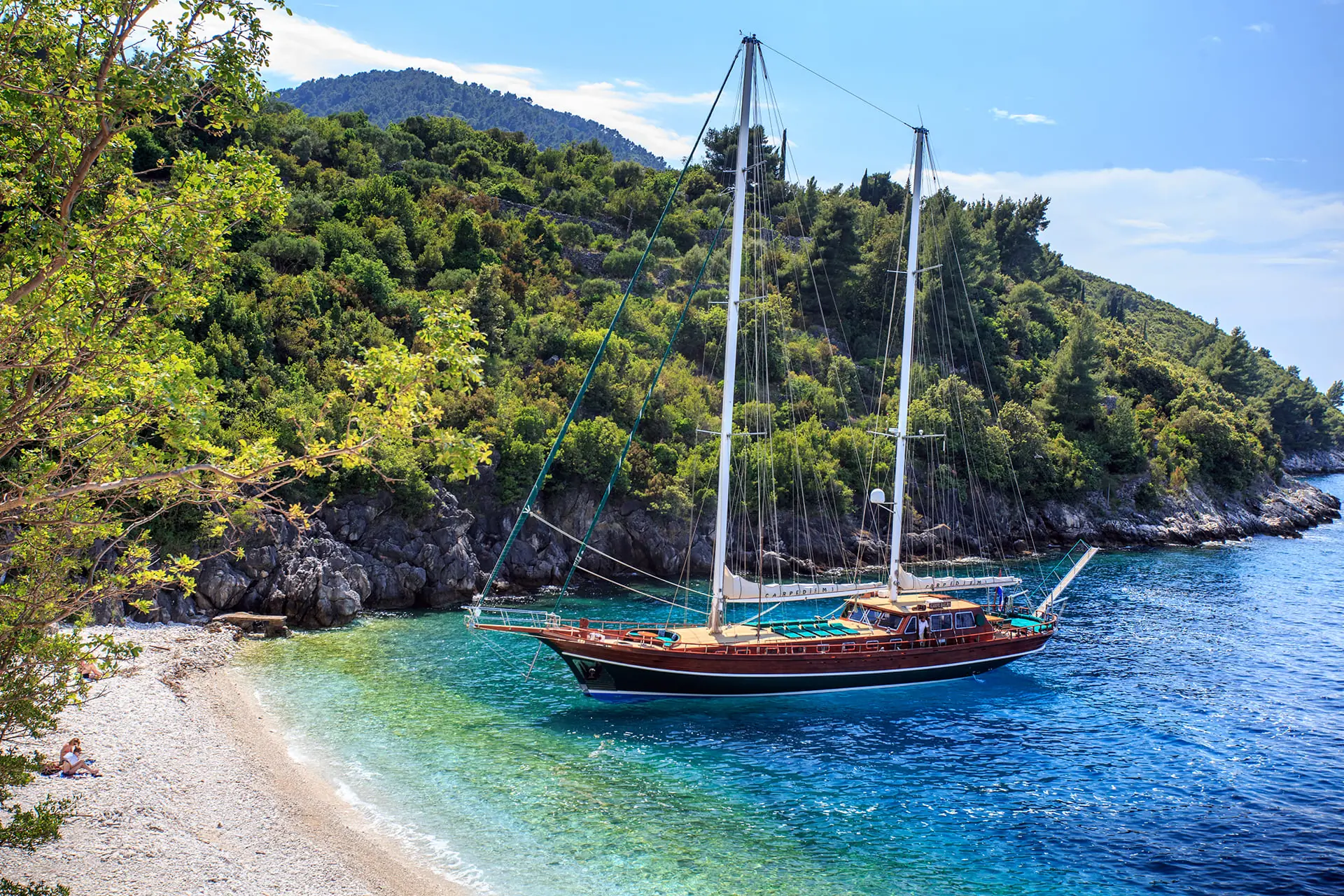 A sailboat with two masts is anchored near a rocky, tree-covered coastline beside a small, sandy beach. The clear blue water fades into turquoise near the shore.