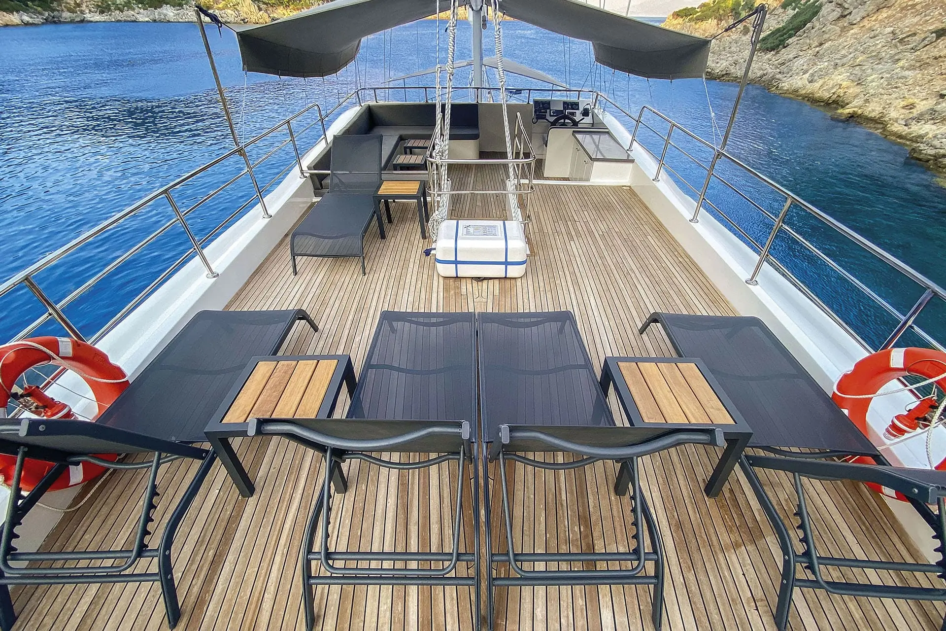 A yacht deck with four lounge chairs, small tables, life preservers, and shaded seating at the back, docked by rocky cliffs and calm blue water.