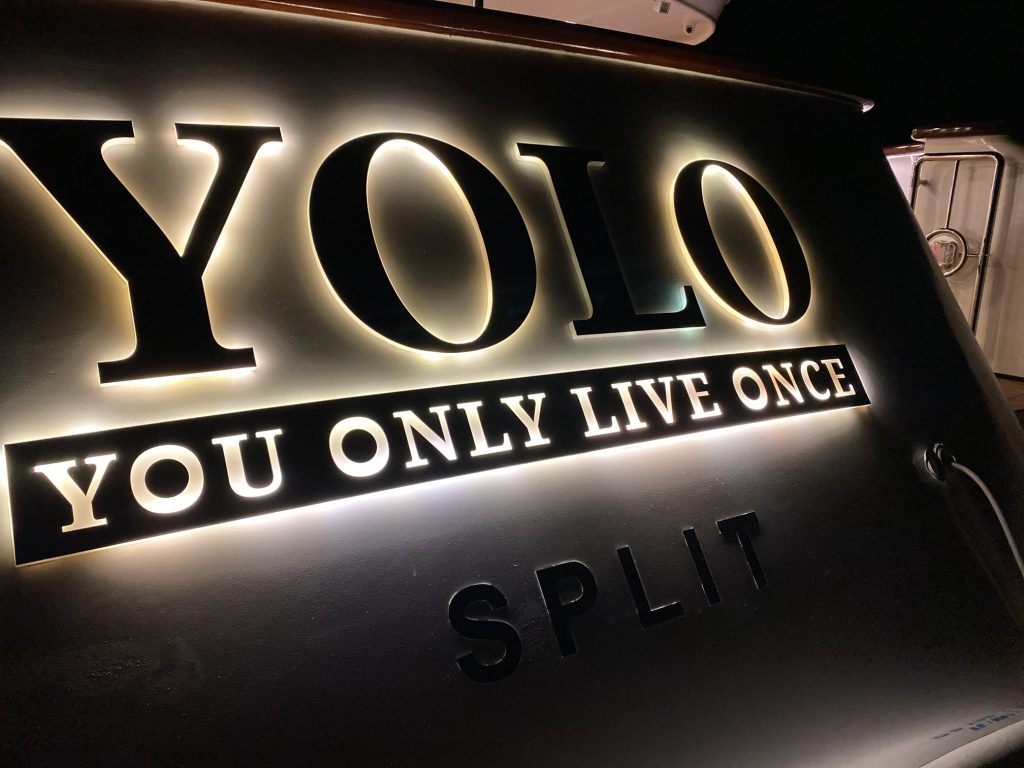 Illuminated letters on a dark surface spell out YOLO and below it, YOU ONLY LIVE ONCE, with SPLIT written underneath, possibly indicating the name and home port of a boat.