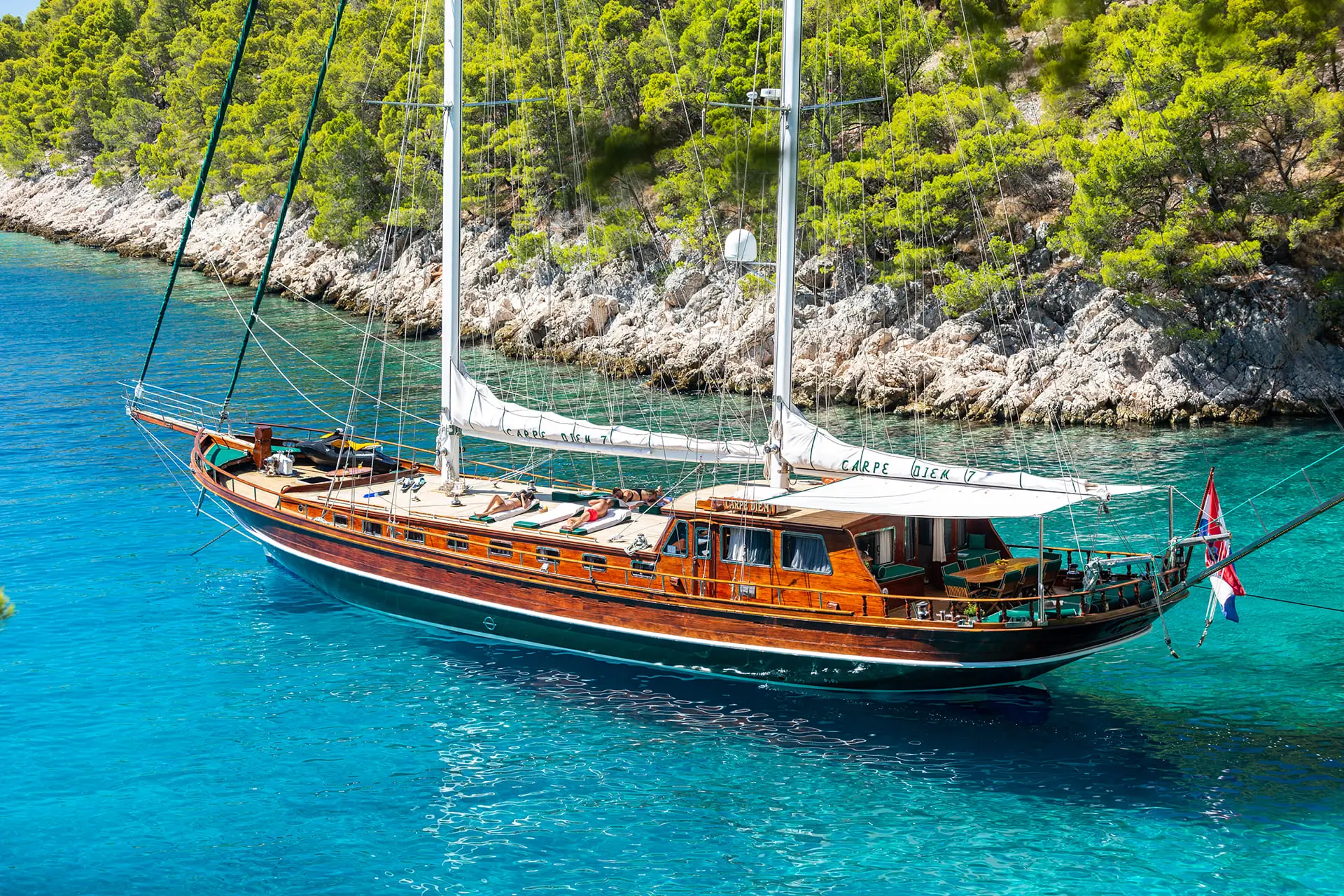 A wooden sailboat with two masts floats on clear blue water near a rocky, tree-covered shoreline. The boats name, CAFFE DICK, is visible on the side.