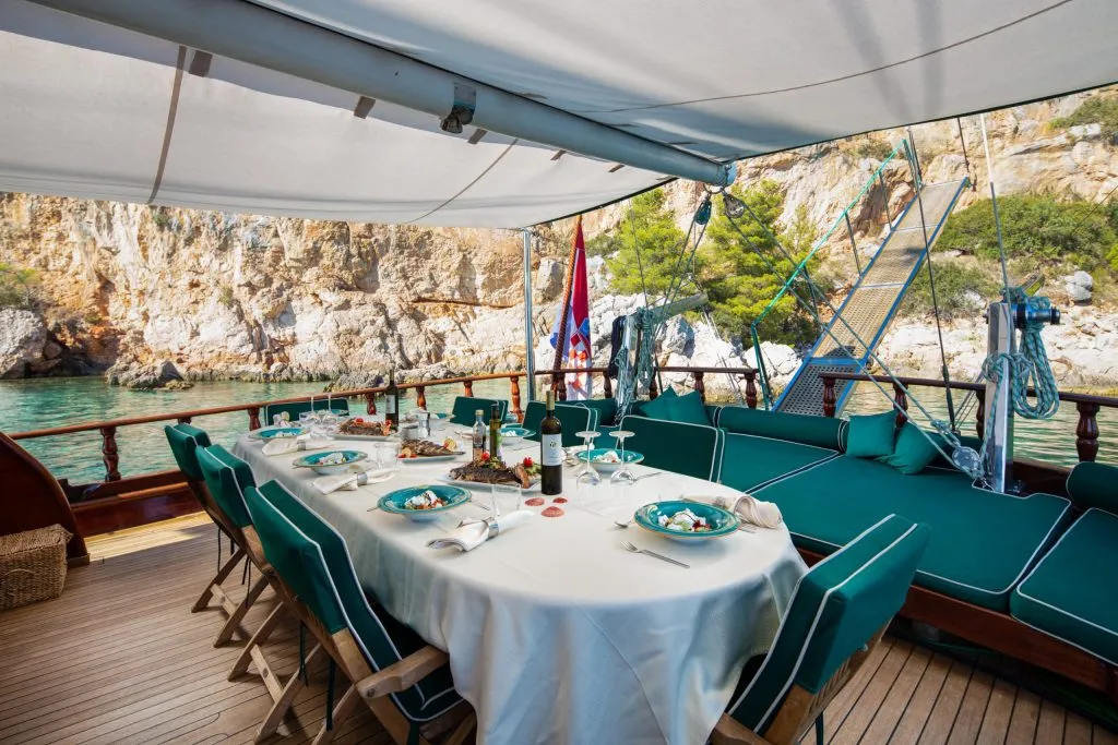 CARPE DIEM 7 A dining table set with plates, glasses, and food sits on the deck of a yacht, shaded by a canopy. Green cushioned seating is nearby, with rocky cliffs and clear water visible in the background.