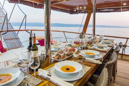 ALTAIR A beautifully set dining table on a yacht, featuring elegant place settings, wine bottles, glasses, and assorted appetizers, with an ocean view and distant hills visible in the background.