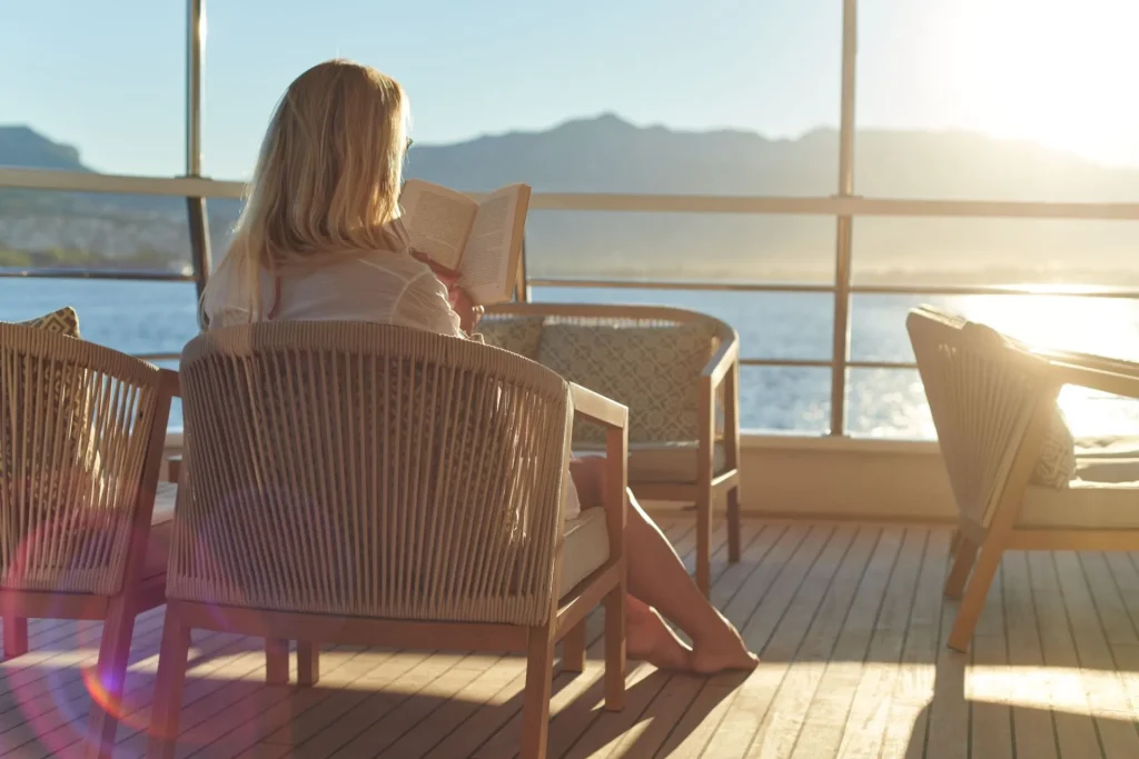 A woman sits on a sunlit deck, reading a book in a wicker chair, with mountains and water visible through large windows in the background. The scene is calm and peaceful, with sunlight streaming in.