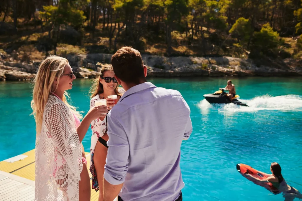 Three people stand on a dock by clear blue water, smiling and holding drinks. In the background, a person rides a jet ski and another floats on an orange raft. Trees and rocks line the shore.
