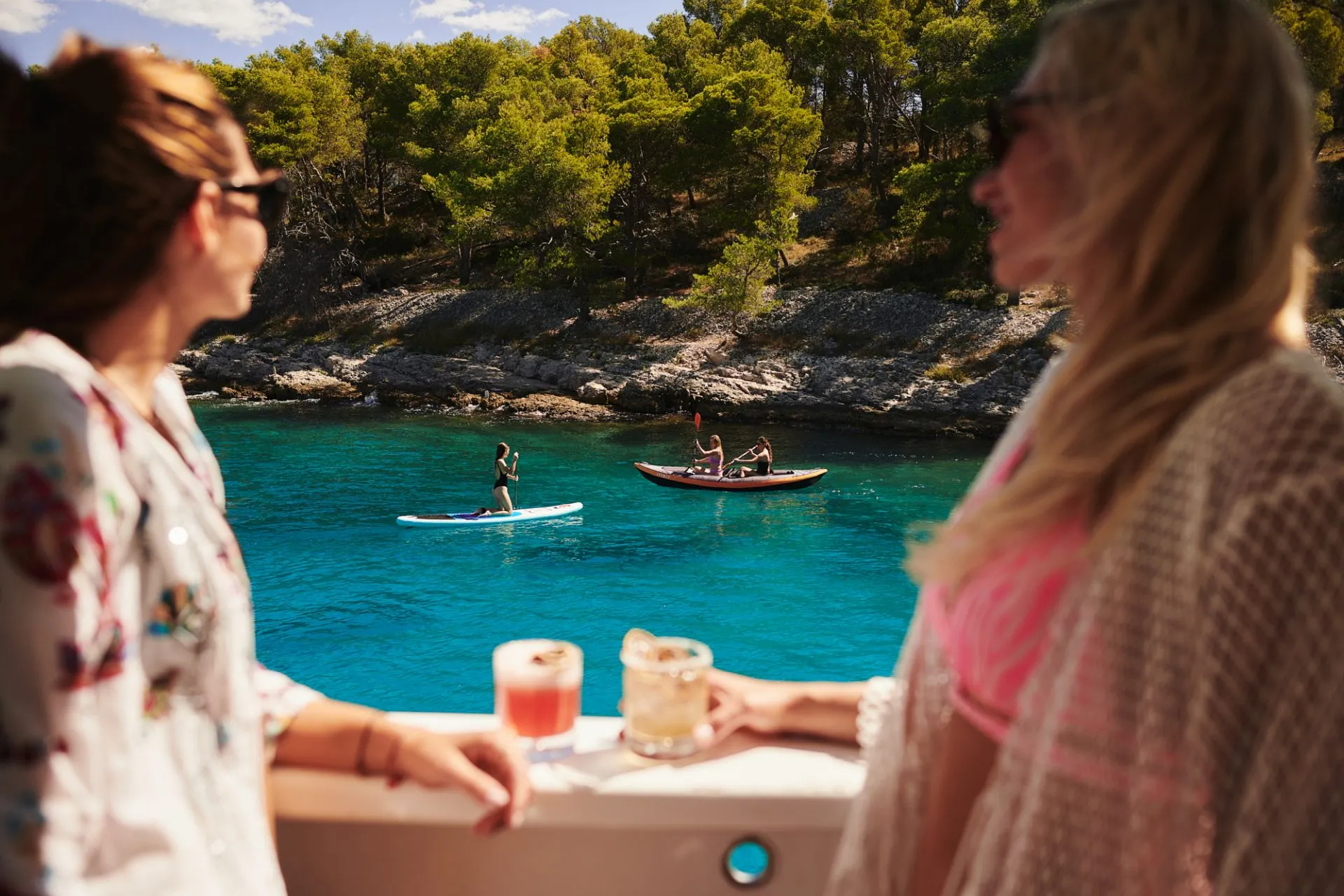 Two women on a boat holding drinks look out at people paddleboarding and kayaking on clear blue water, with a forested shoreline in the background.
