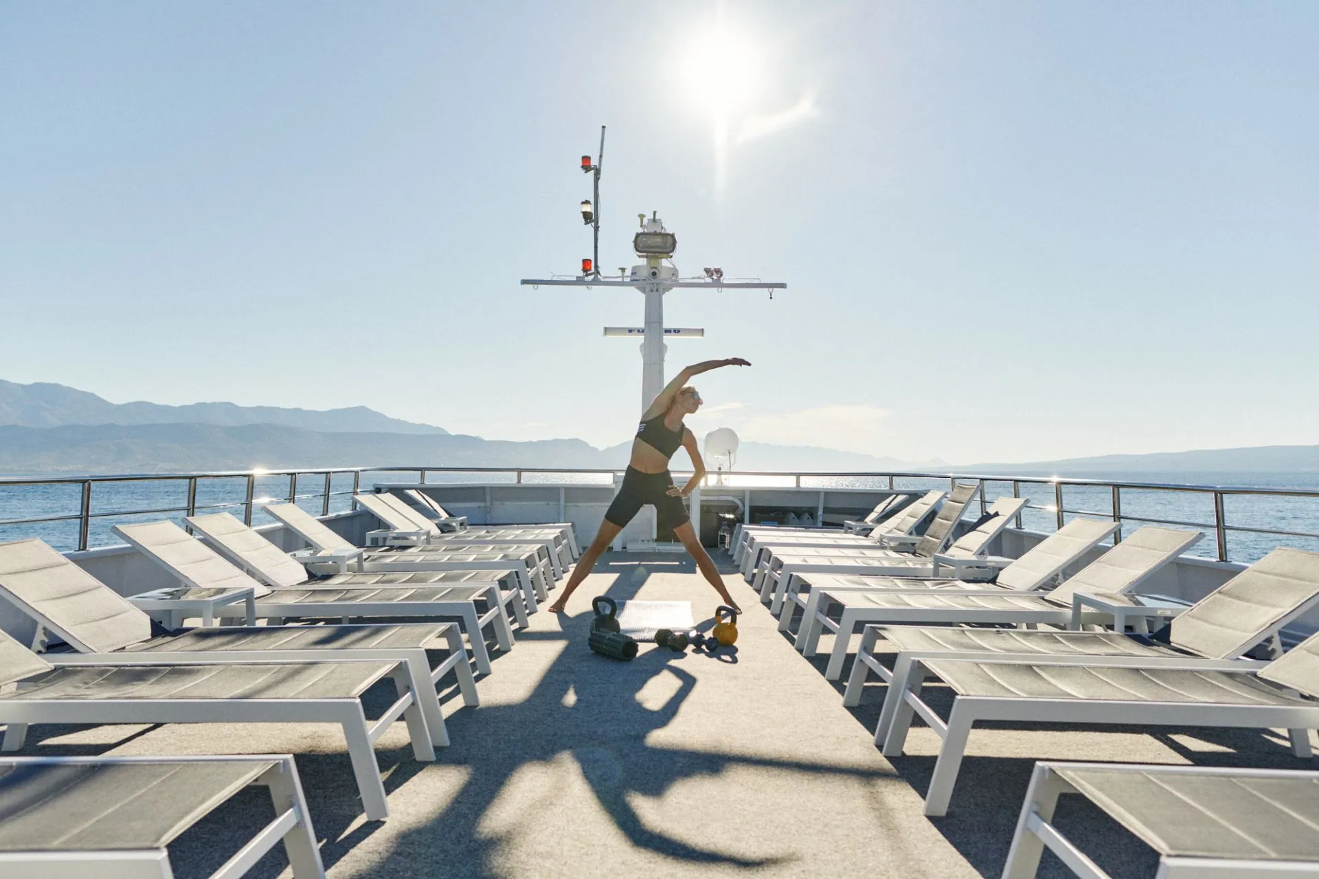 A person in athletic wear stretches on the deck of a boat, surrounded by empty lounge chairs, with kettlebells nearby. The sun is shining brightly, and calm water and distant mountains are visible in the background.