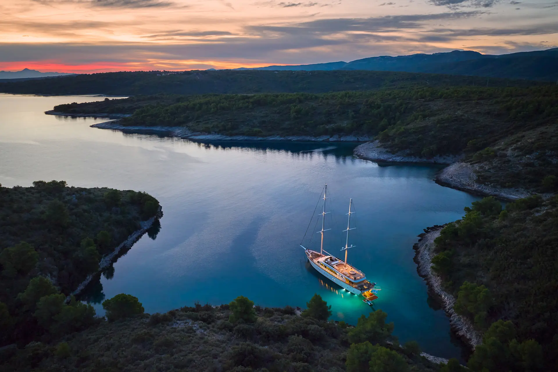 A yacht with glowing underwater lights is anchored in a quiet bay surrounded by forested hills at sunset, with calm water reflecting the colorful sky.