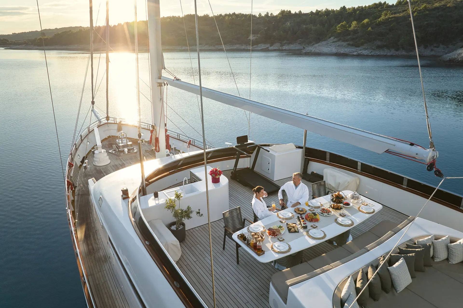 Two people sit at a beautifully set dining table on the deck of a yacht, enjoying a meal at sunset on calm water, with a scenic forested shoreline in the background.