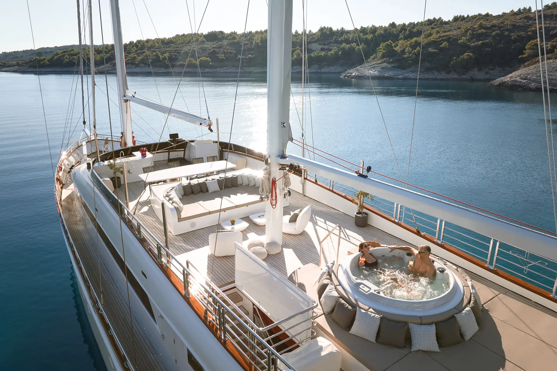 A luxury yacht deck features a hot tub with two people relaxing, surrounded by lounge seating and calm blue water with a forested shoreline in the background.