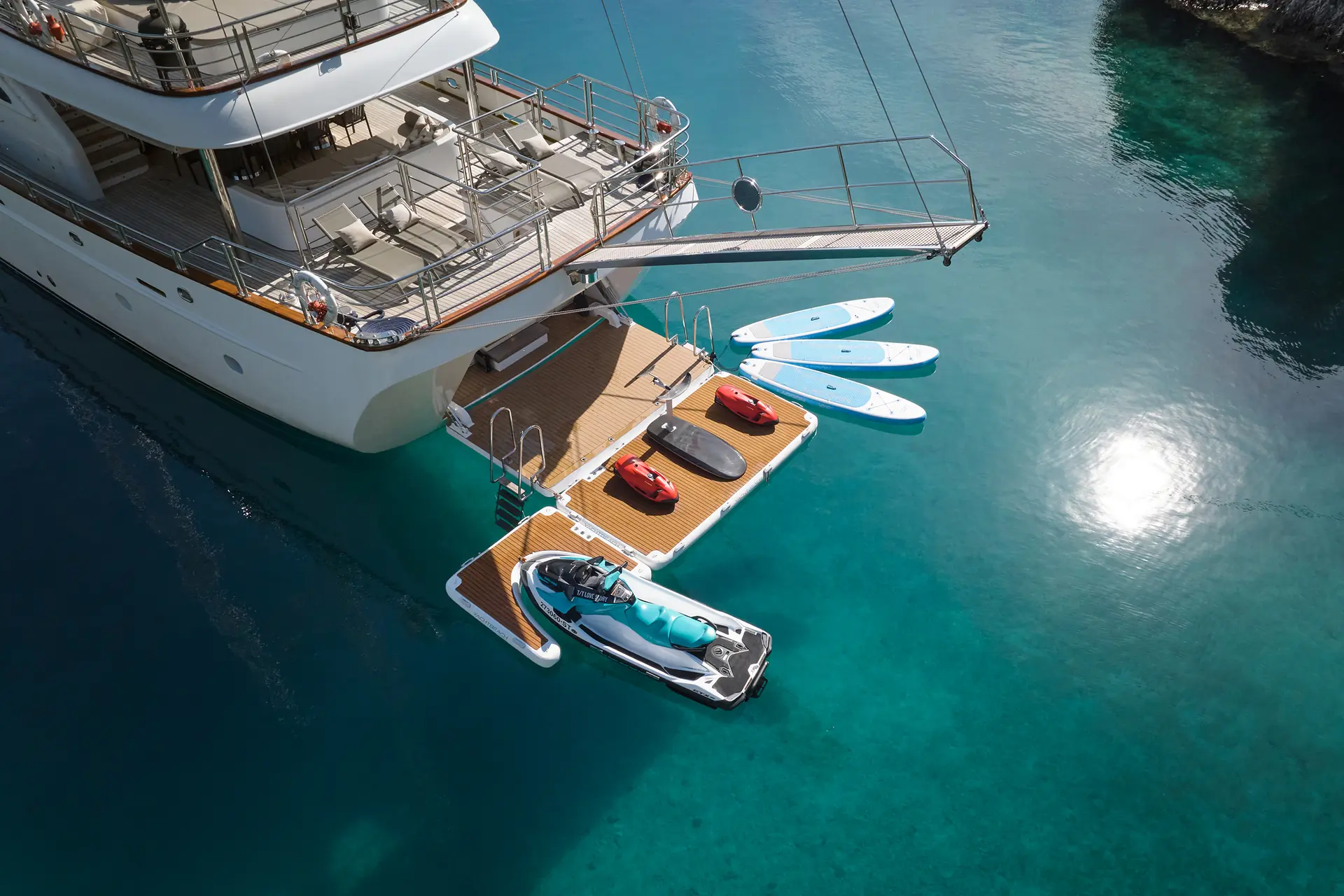 Luxury yacht docked on calm, clear blue water with a floating platform holding a jet ski, kayaks, and paddleboards. Sunlight reflects off the water, creating a serene, inviting atmosphere for water activities.