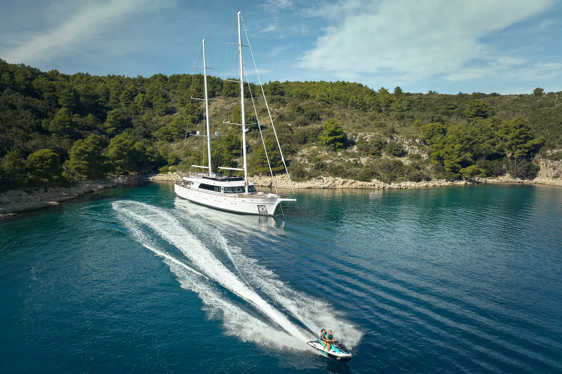 A white yacht is anchored in a calm, clear blue bay near a forested shoreline, while two people ride a jet ski, creating a trail in the water beside the yacht.