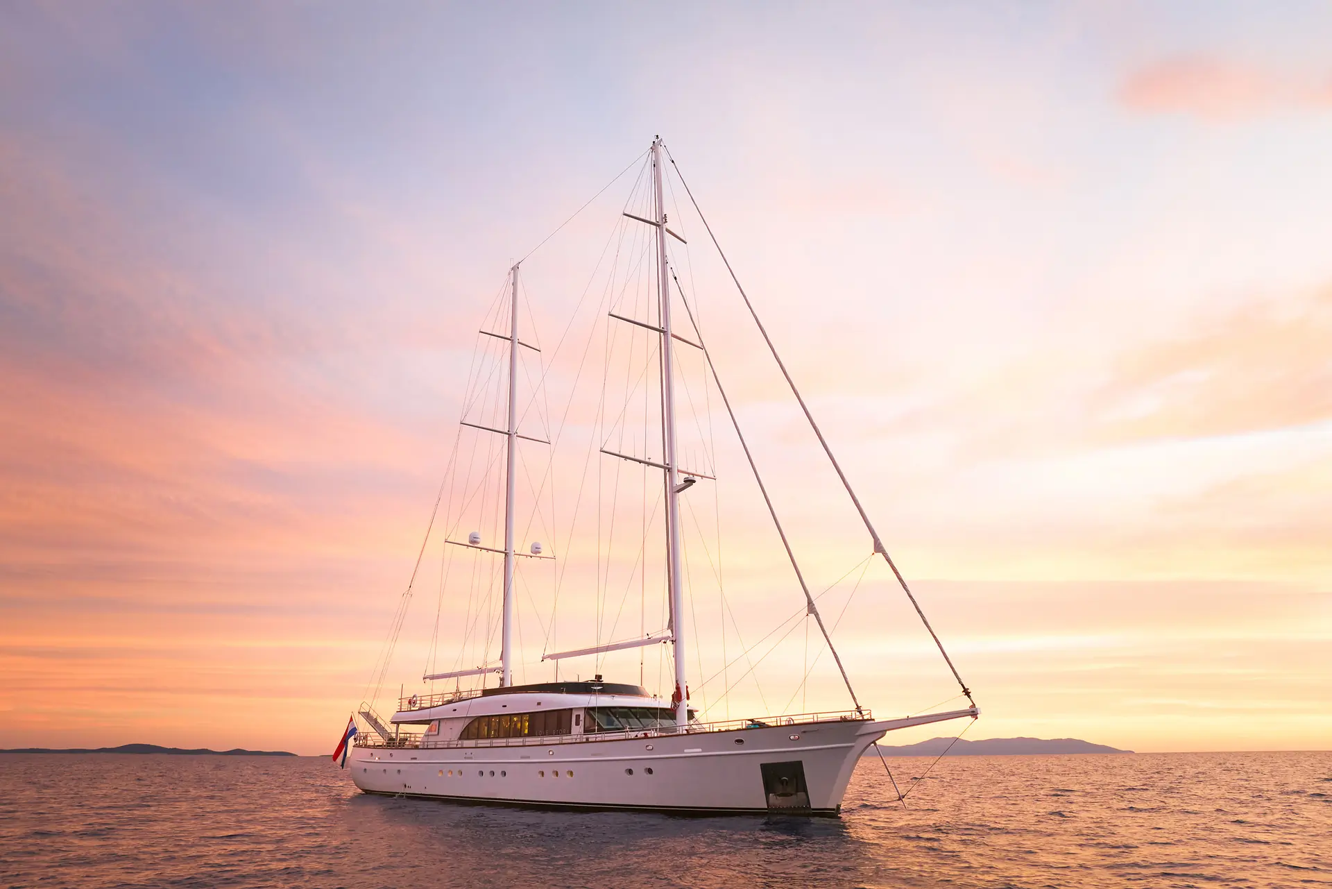 A large white sailboat floats on calm water during a pastel-colored sunset, with soft pink and orange clouds in the sky and distant land visible on the horizon.