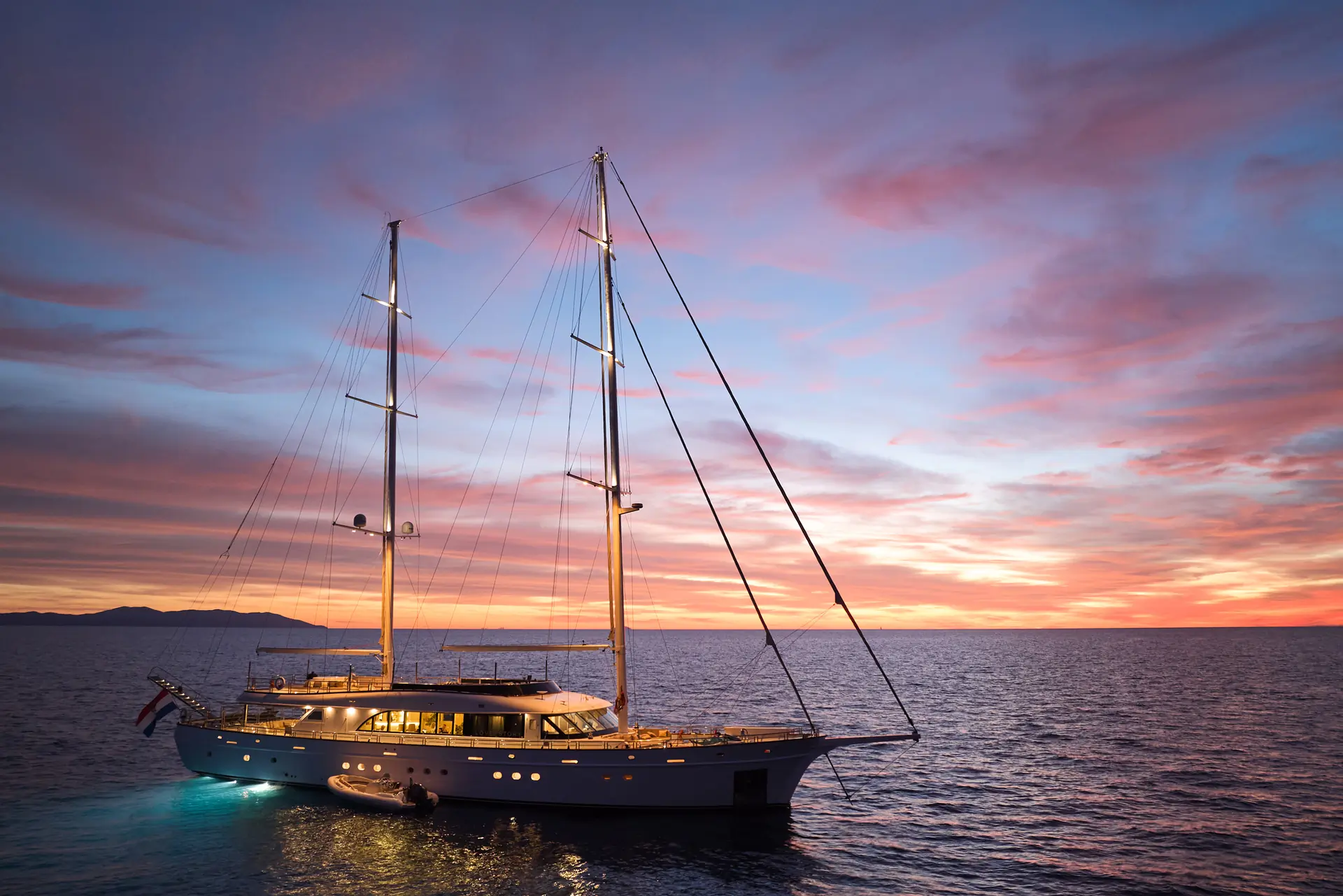 A large sailboat with glowing lights floats on calm water at sunset, with a colorful sky and distant land on the horizon.