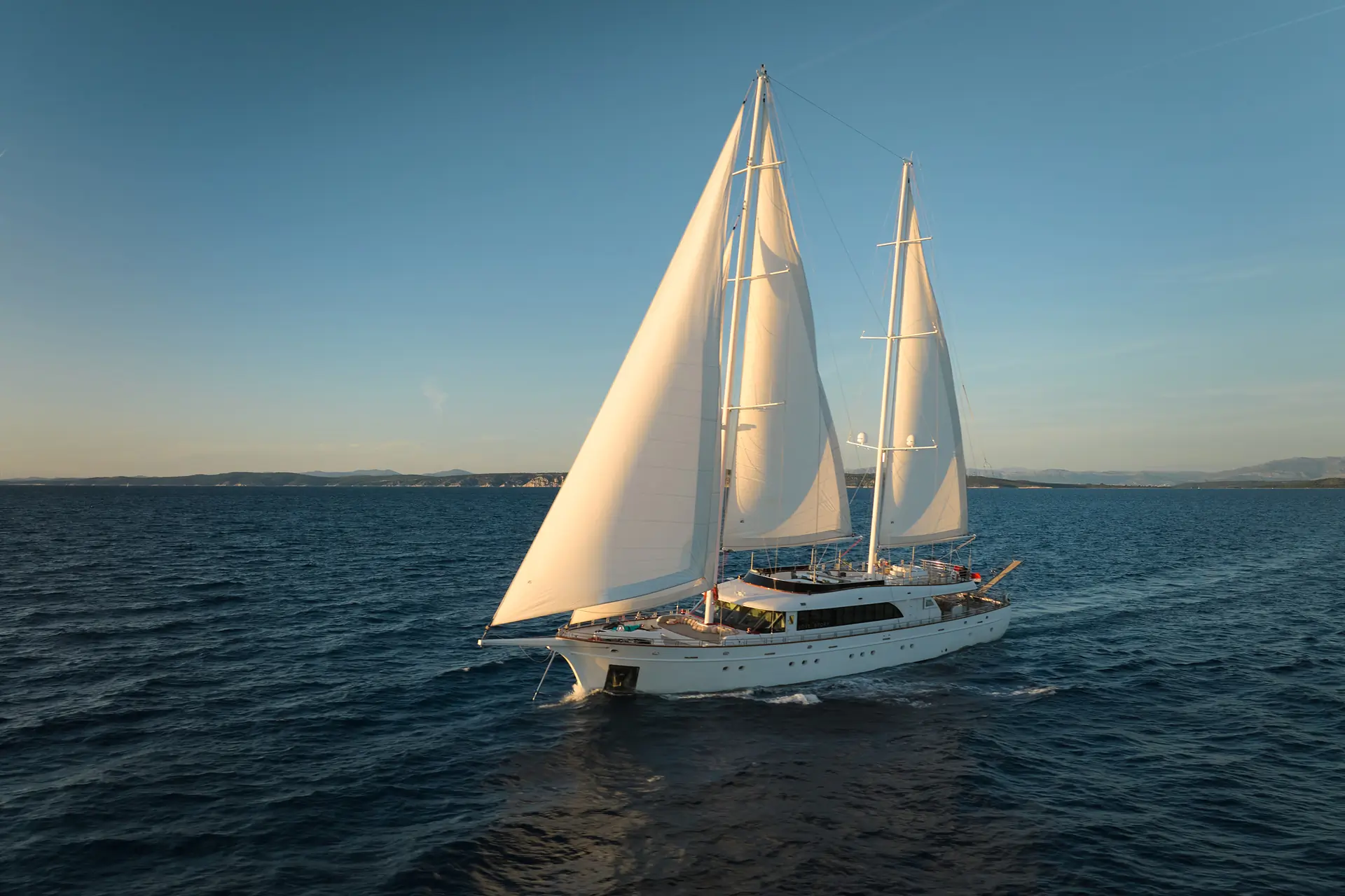 A large white yacht with two sails glides on calm blue water under a clear sky, with distant coastline and low hills visible in the background.