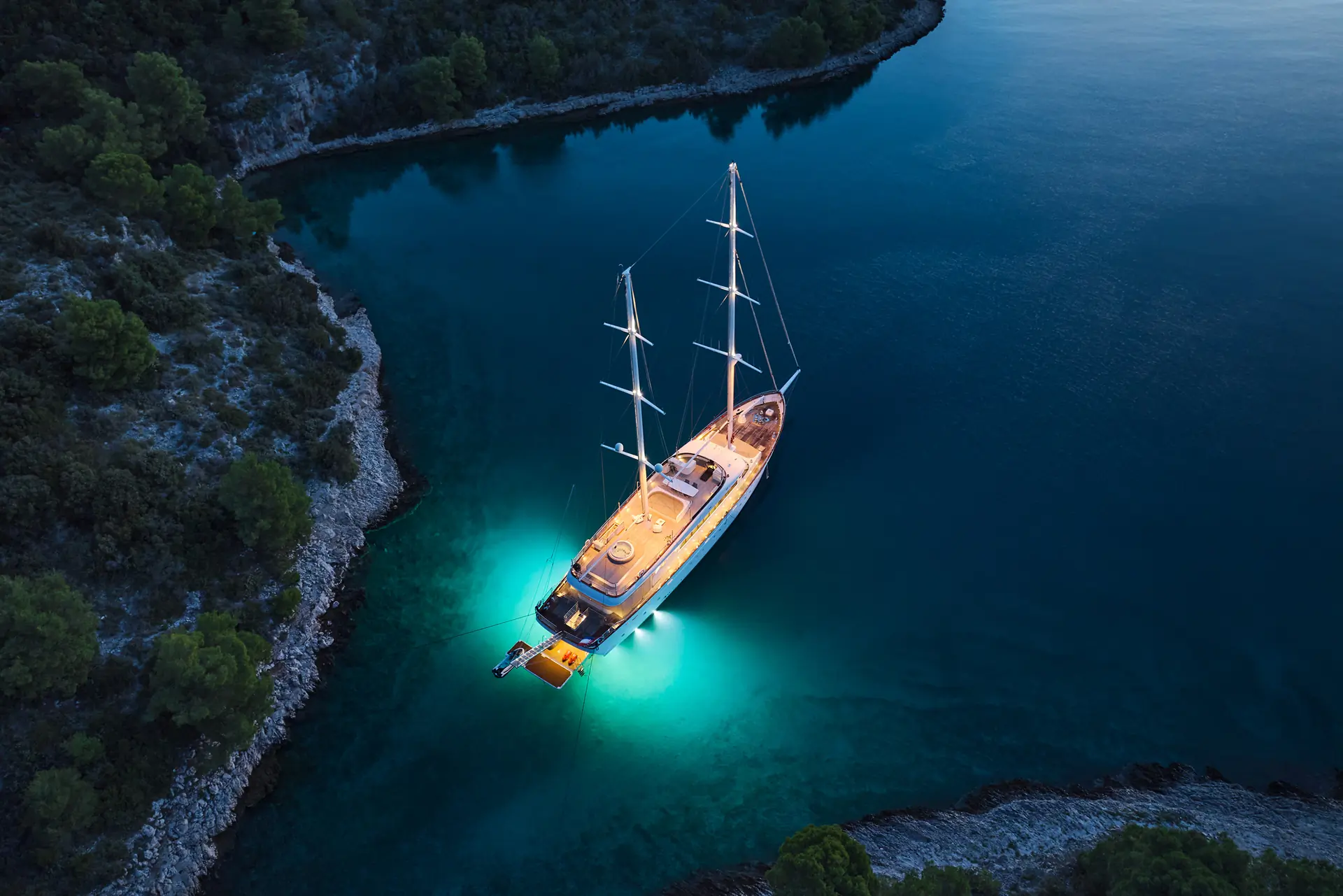 A yacht with deck lights anchored in a calm, narrow bay at dusk, surrounded by forested hills and rocky shoreline, with glowing water beneath the boat.