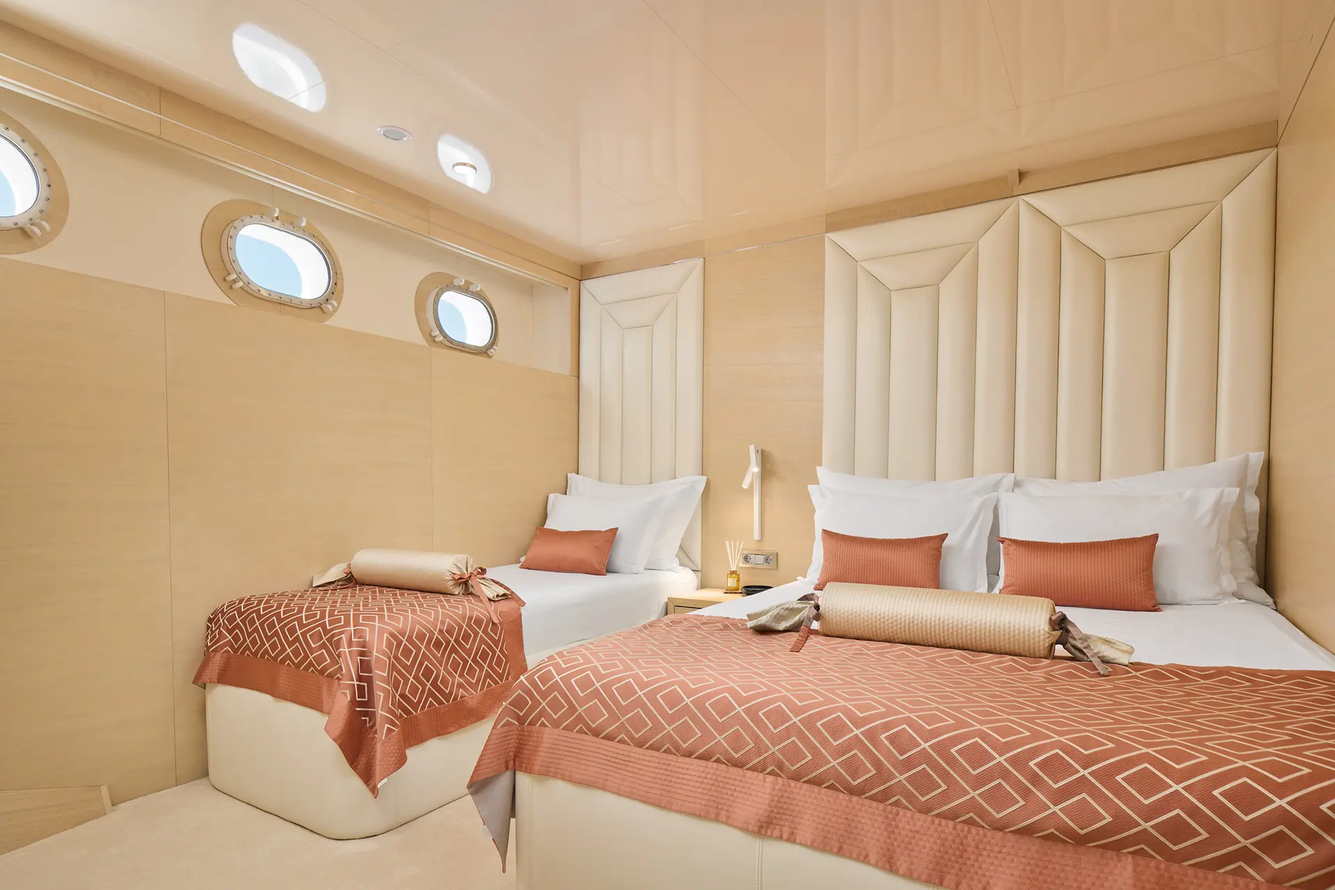 A modern yacht cabin with two beds, beige walls, geometric headboards, three round porthole windows, and rust-colored bedding with matching pillows. A small lamp sits on a nightstand between the beds.