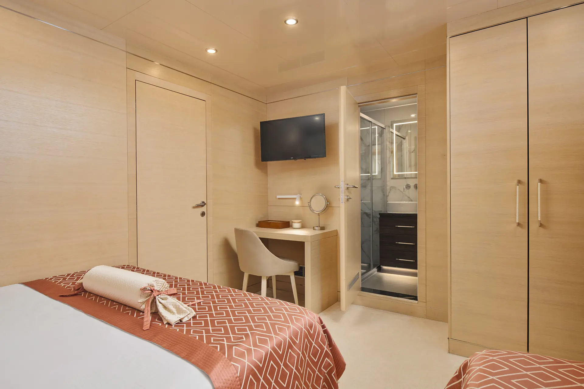 A modern bedroom with two single beds, a desk with a chair and mirror, a wall-mounted TV, an open door leading to a bathroom, and light wood furniture and walls.