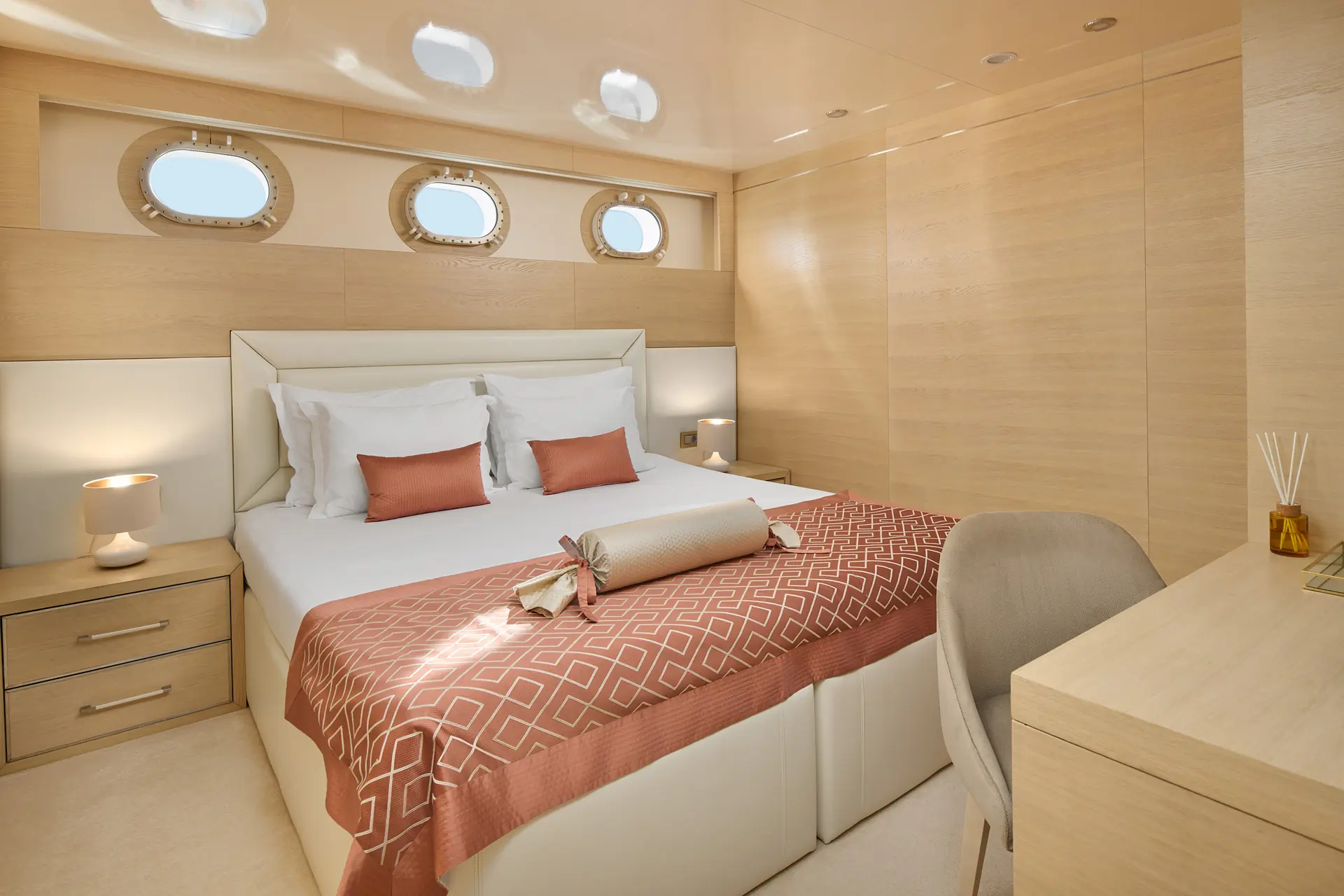 A modern yacht bedroom with a double bed, peach bedding, three round portholes, nightstands with lamps, a chair, desk, and light wood walls and furniture.