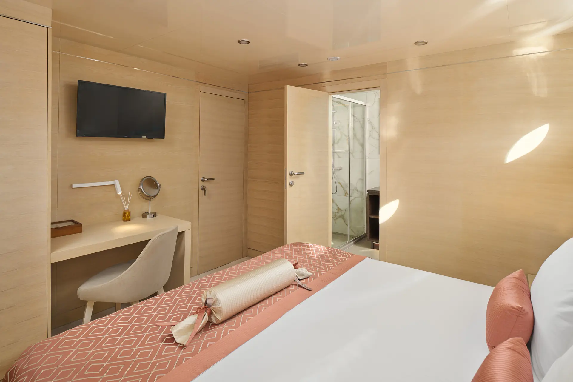 A modern bedroom with a bed, pastel pink bedding, a desk with a chair and lamp, a wall-mounted TV, and an open door revealing a marble bathroom. Light wood walls and natural light create a bright, cozy space.