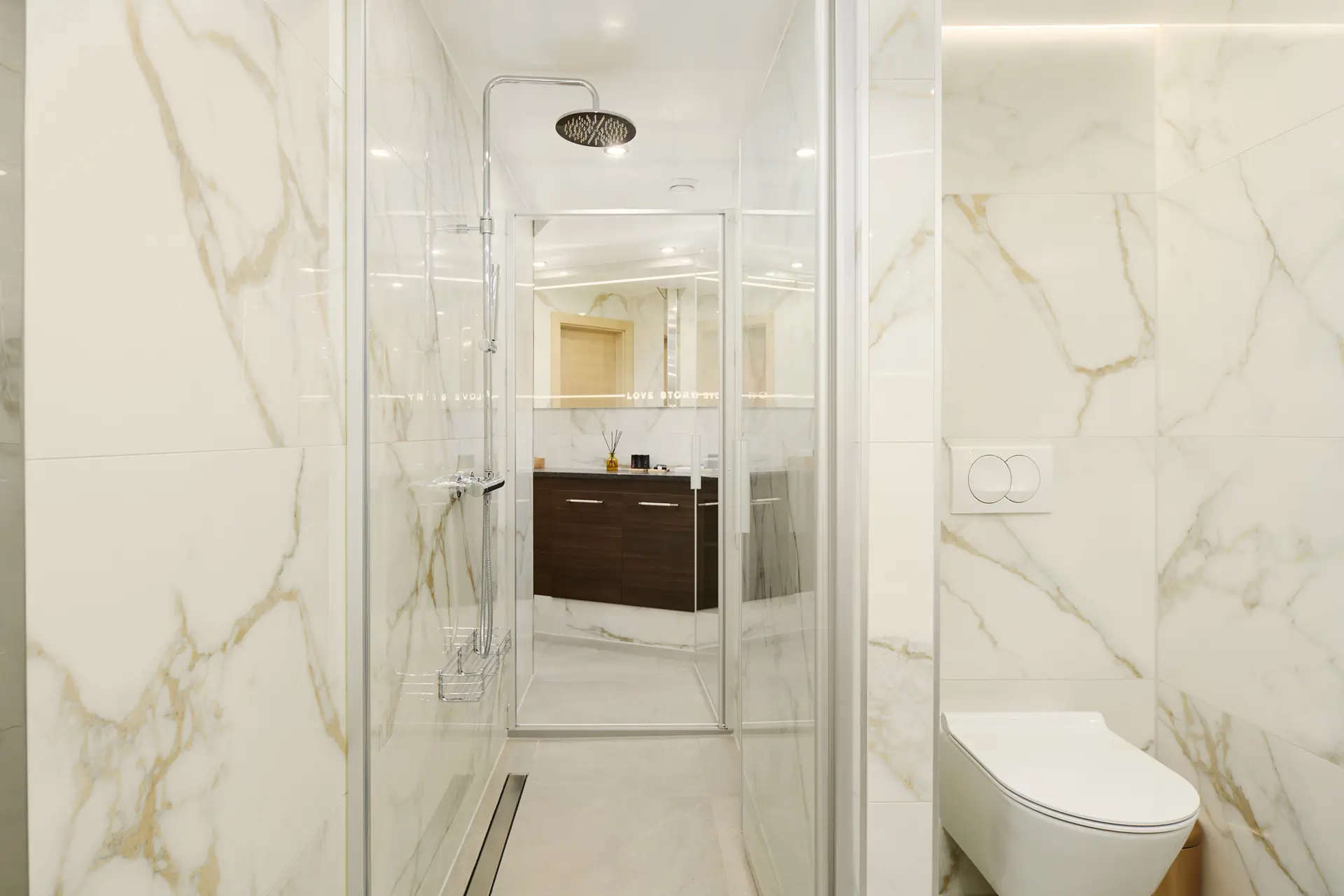Modern bathroom with marble walls, a glass-enclosed shower with a rainfall showerhead, a wall-mounted toilet, and a vanity with a mirror in the background.
