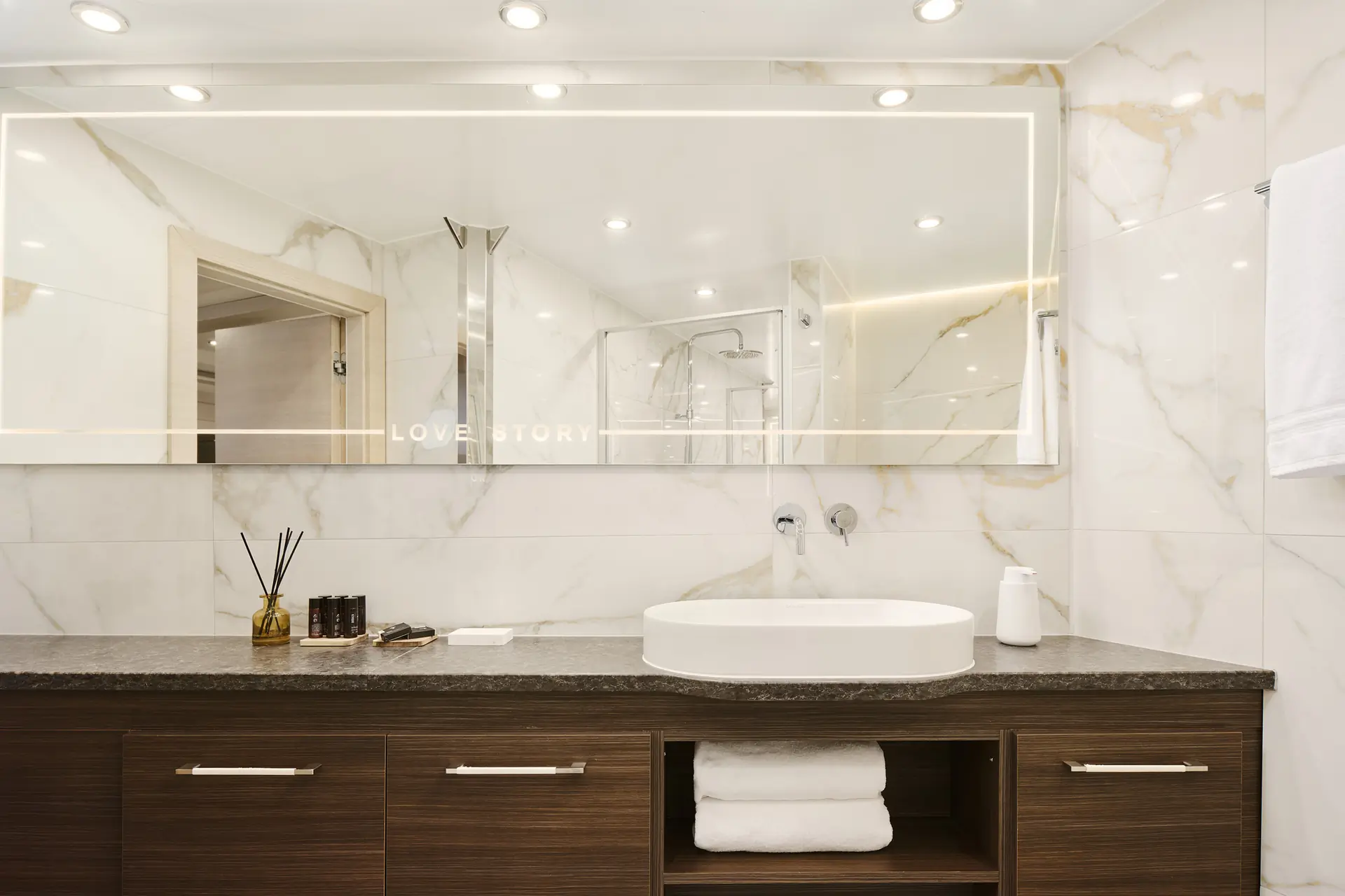Modern bathroom with a large mirror, white sink, dark countertop, wooden cabinets, folded towels, toiletries, and marble-patterned walls, all brightly lit by recessed ceiling lights.