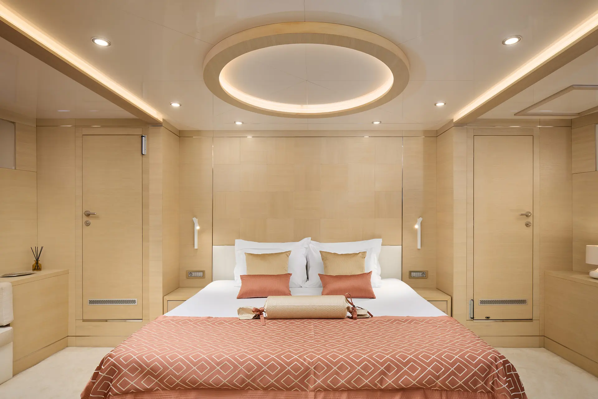 Modern bedroom with beige walls and ceiling, circular recessed lighting, a large bed with white linens and peach pillows, matching peach blanket, and minimalist decor. Two wooden doors and built-in lights are visible.