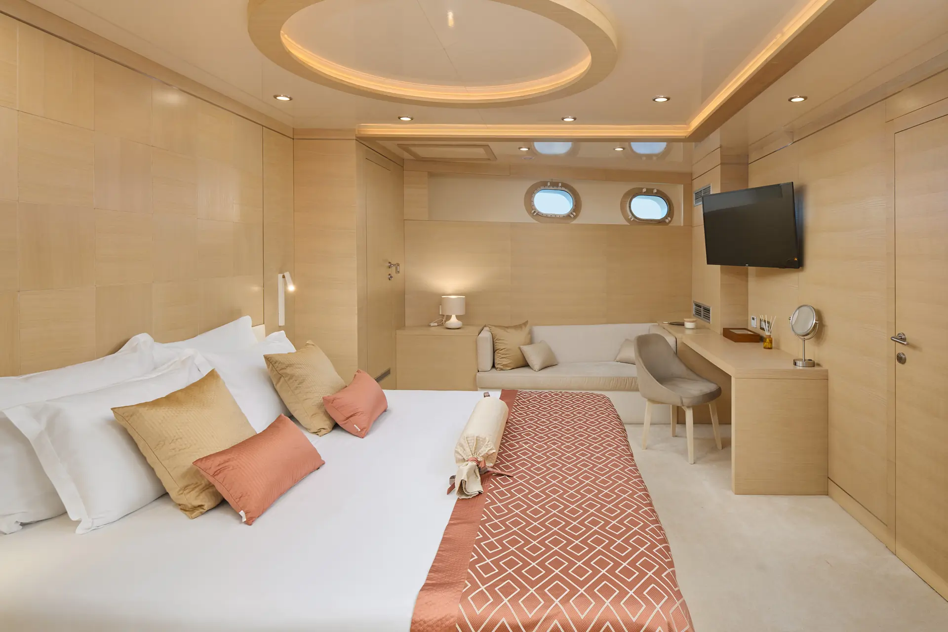 A modern yacht bedroom with a large bed, beige walls, tan and coral accents, a wall-mounted TV, small desk, sofa, and porthole windows. The space feels bright, cozy, and luxurious.