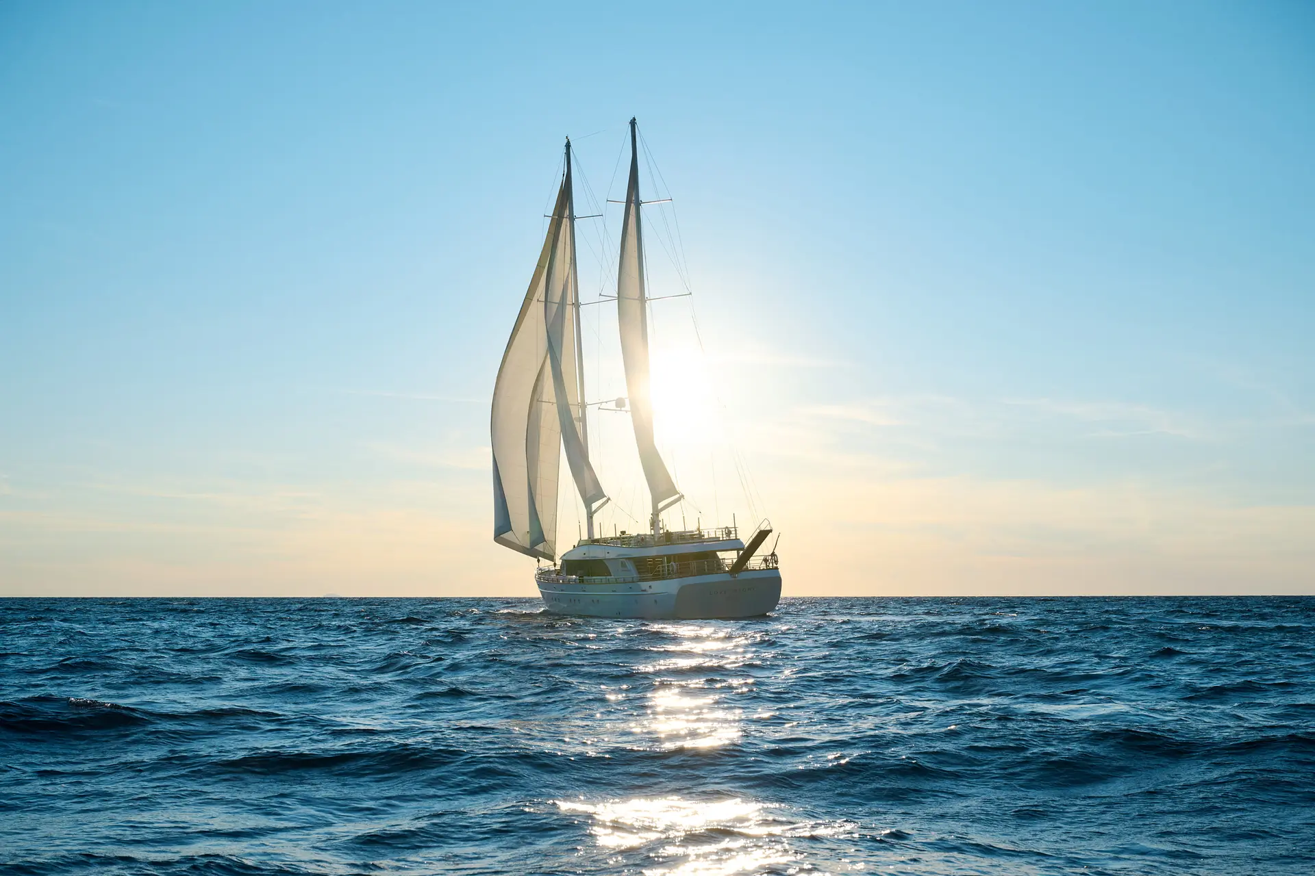 A sailboat with two sails glides on the ocean at sunset, with sunlight shining through the sails and reflecting on the waters surface.