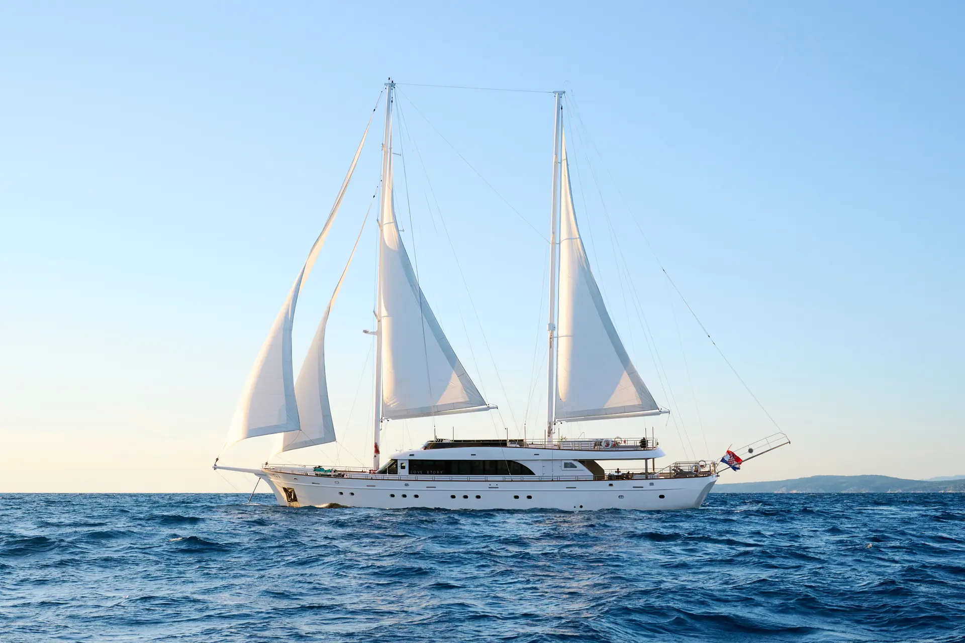 A large white sailing yacht with two masts sails on calm blue sea under a clear sky. The yacht has several sails unfurled and a flag at the stern. Land is visible in the distance on the horizon.