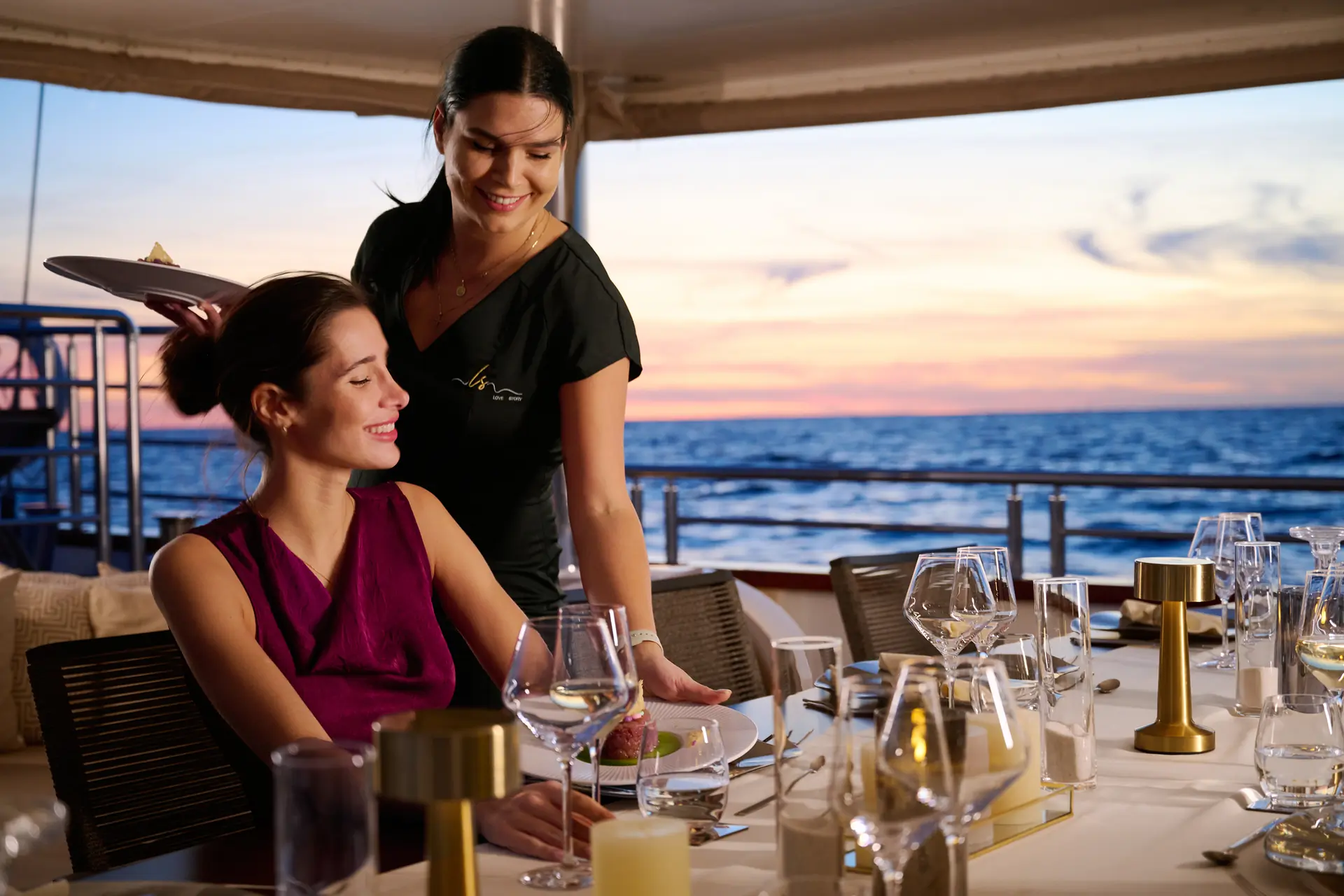 A smiling server brings dessert to a seated woman at an elegant, candlelit dining table on a ships deck, with the ocean and a colorful sunset in the background.