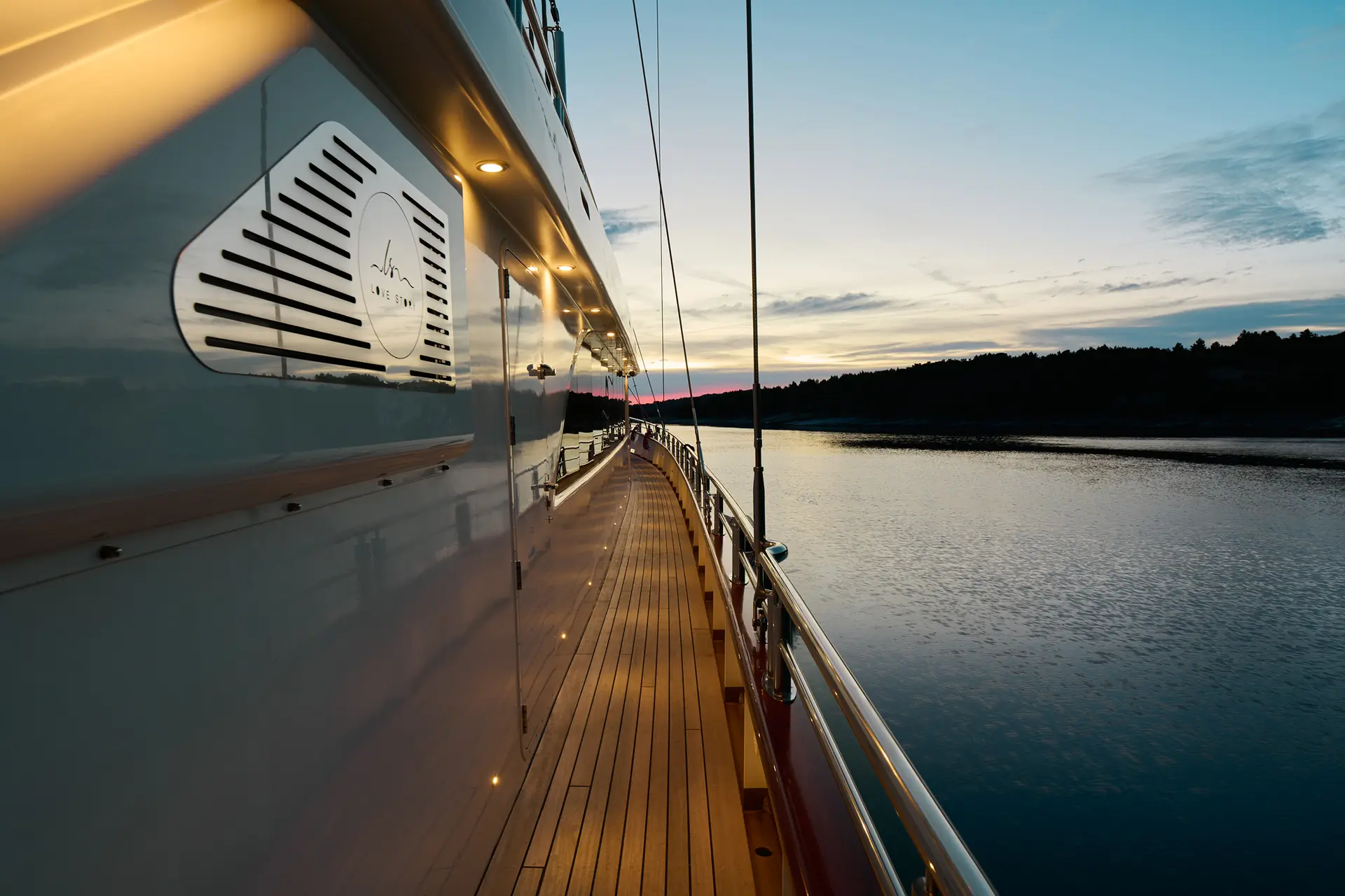 A view along the wooden deck of a yacht at sunset, with calm water and a distant tree-lined shore under a partly cloudy sky.