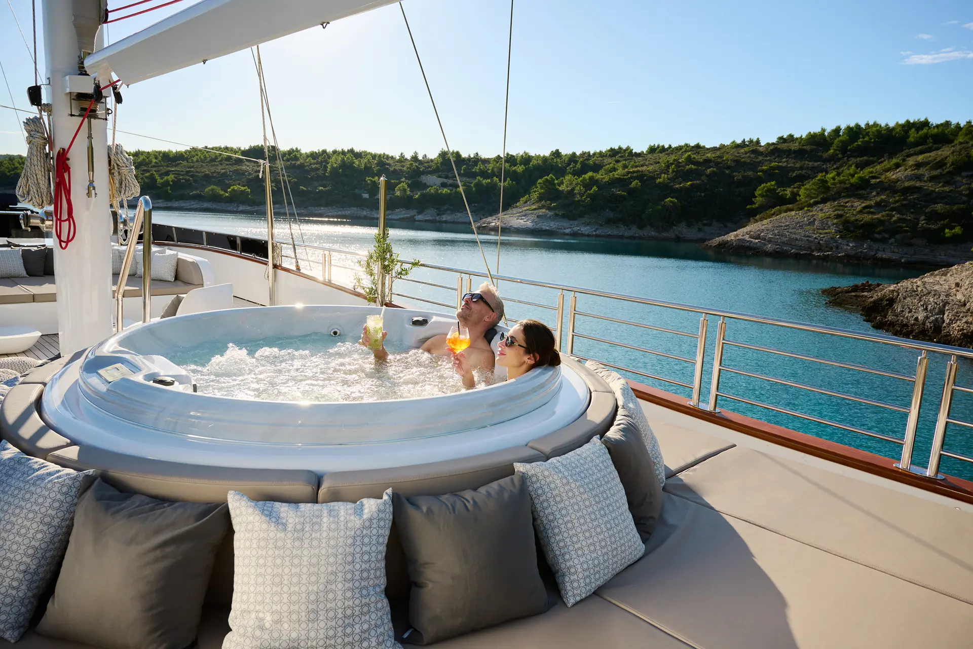 A man and woman relax in a hot tub on a yacht, holding drinks. The yacht is anchored near a scenic, tree-lined shoreline under a clear blue sky, with water and hills in the background. Cushions line the deck nearby.