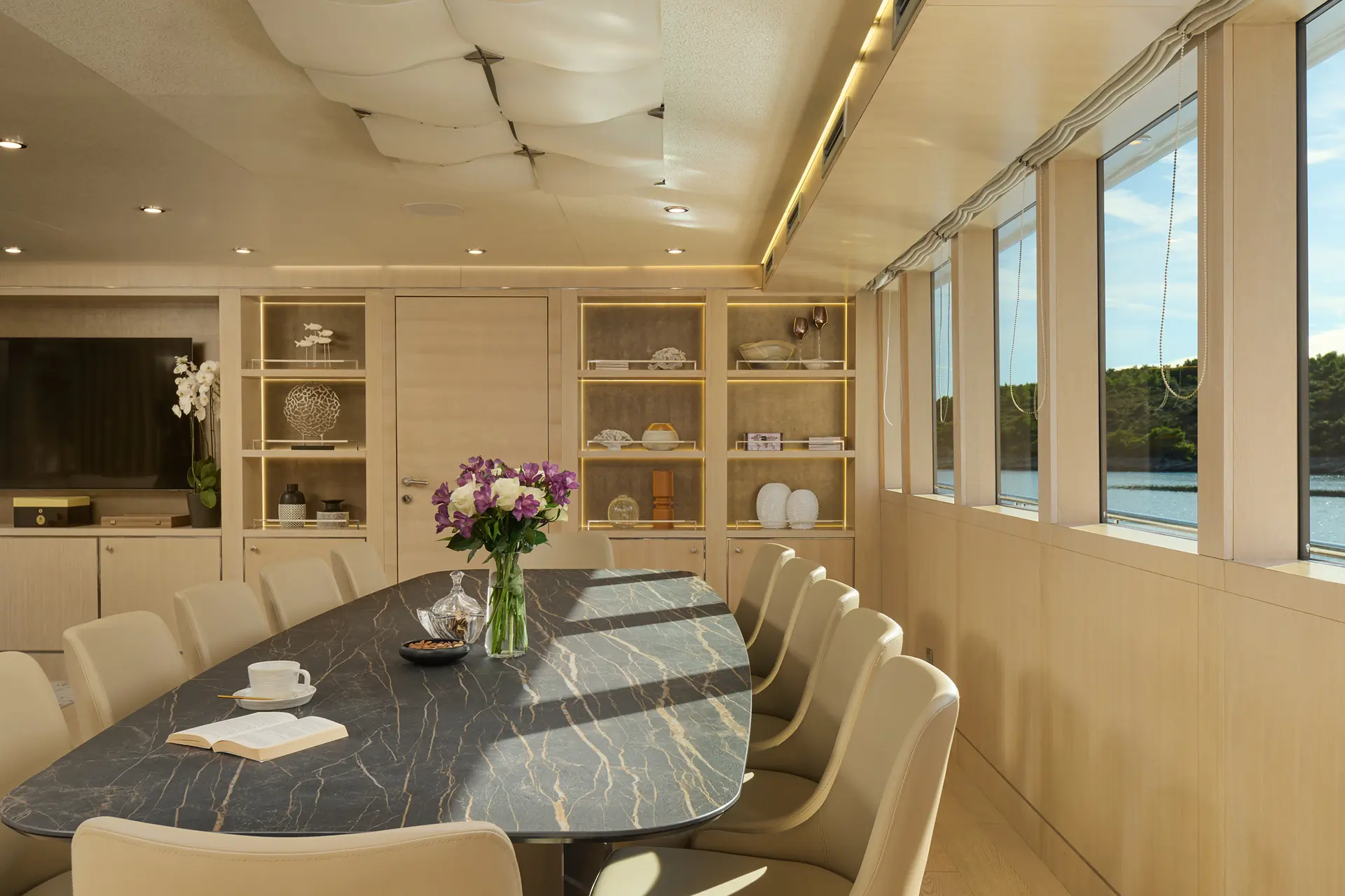 Elegant dining room with a long marble table, beige chairs, and shelves with decor. Large windows on the right show a water view. A vase of purple flowers and an open book are on the table. Warm, modern lighting overhead.