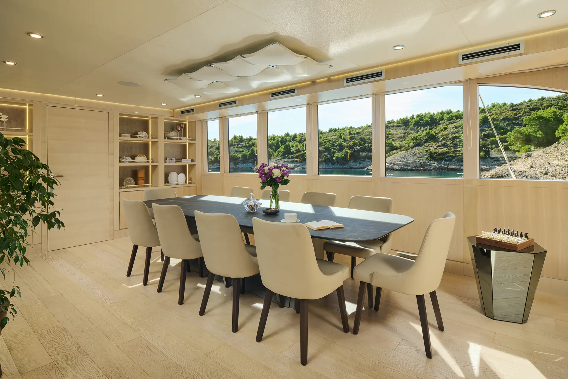 A modern dining room with a dark table set for eight, cream chairs, flowers, books, and large windows showing a scenic water and forest view. A chessboard is on a small side table.