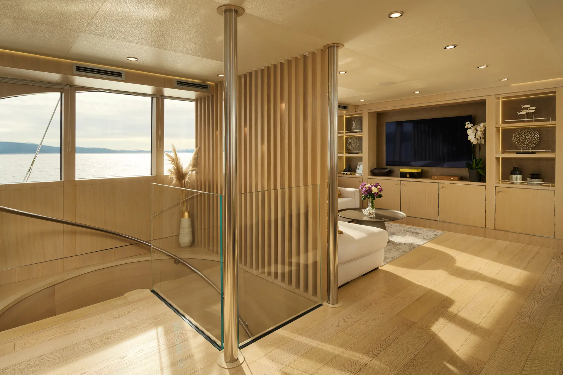 Modern yacht interior with light wood finishes, a spiral staircase, built-in shelves, large windows with an ocean view, a white sofa, a TV, and decorative items including flowers and plants.
